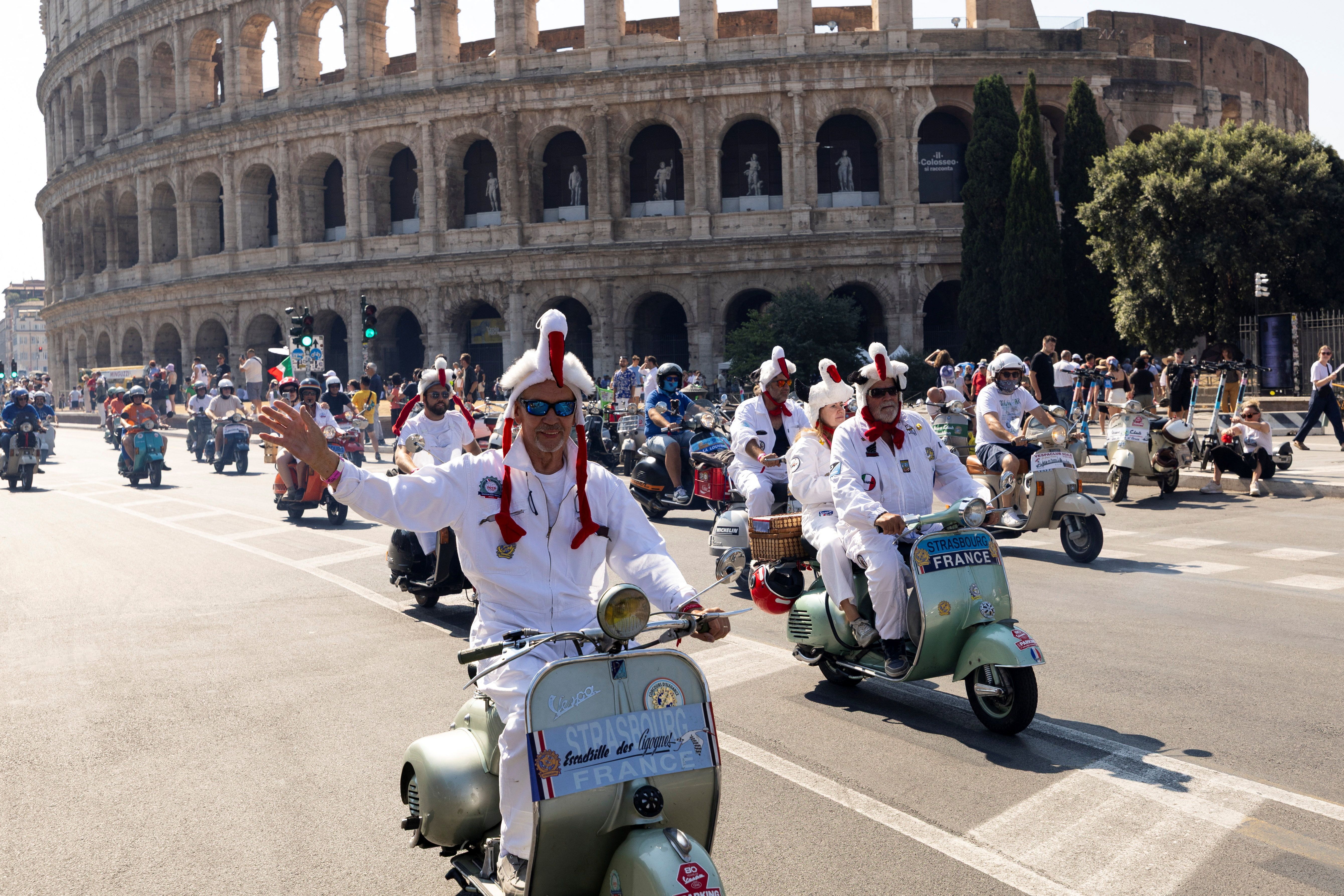 Scooter parade near the Colosseum in Rome: riders dressed in white with headpieces ride vintage scooters as crowds watch; Italian flags flap under the ancient arches.