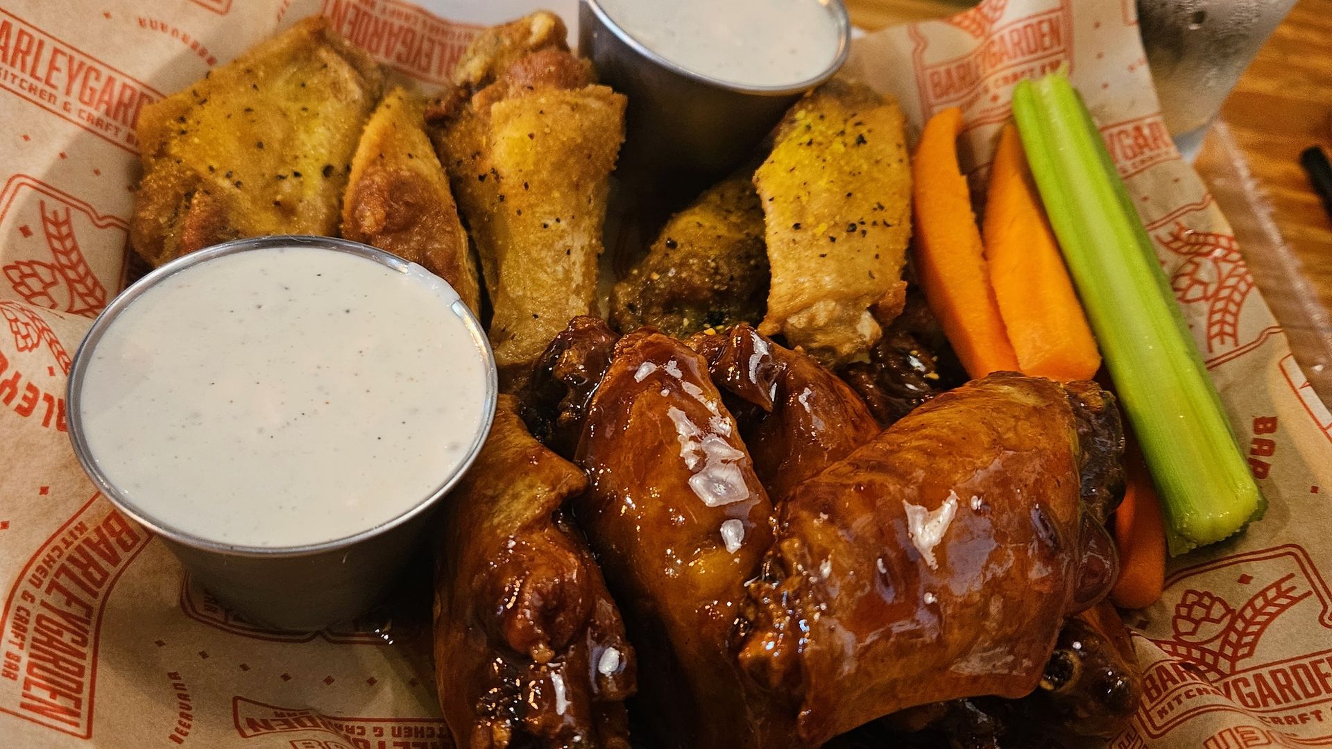 A basket of crispy golden and saucy chicken wings served with ranch dressing, carrot sticks, and celery sticks on Barleygarden-branded paper.