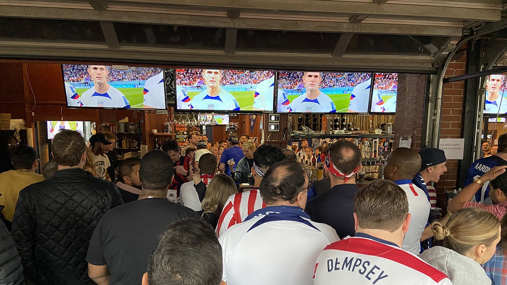 Busy sports bar with four large screens showing a soccer player in a white and blue USA jersey. Crowded patrons wear red, white, and blue; brick walls, bar taps, and busy shelves in the background.