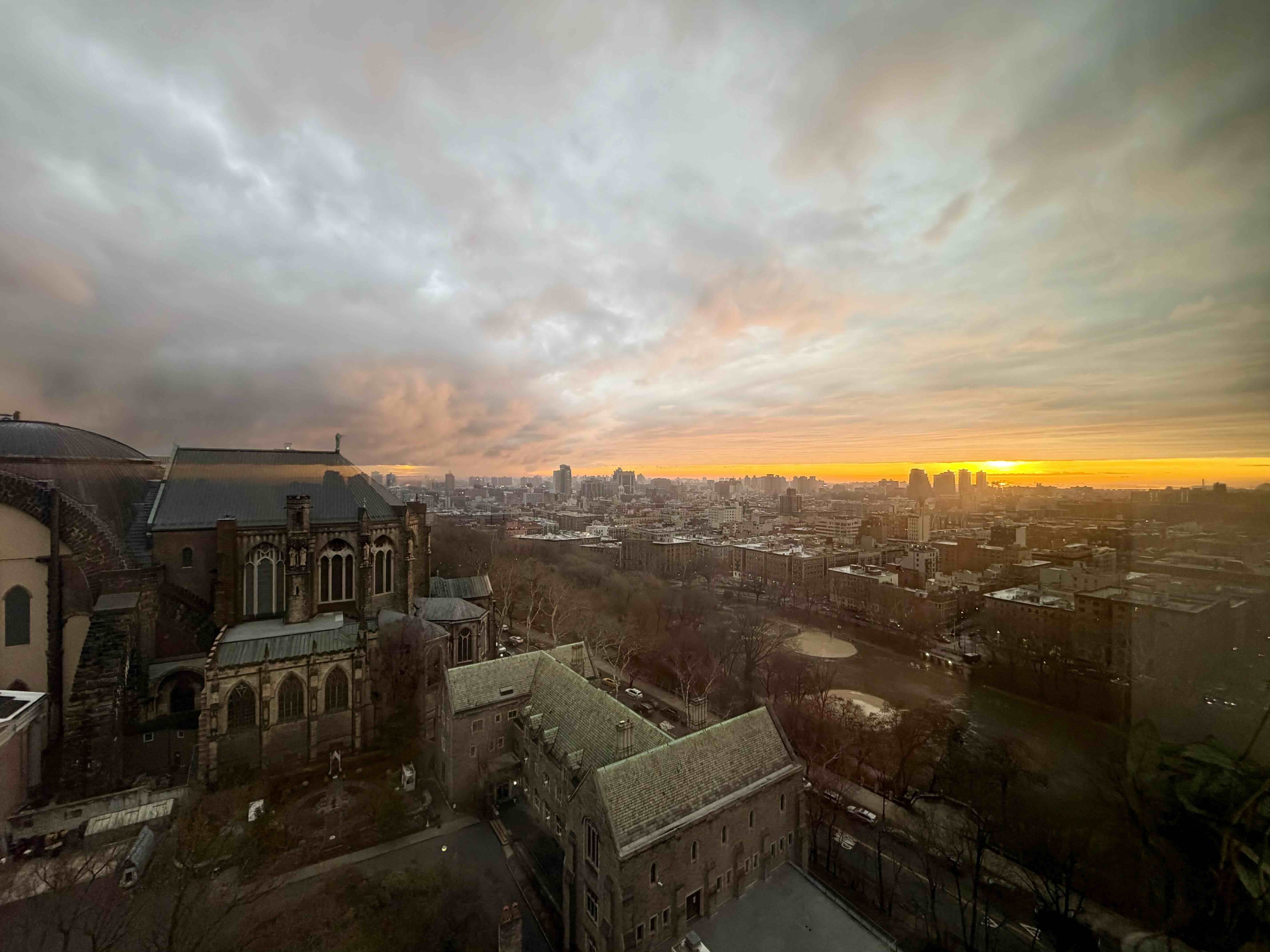 Overlooking a city at sunset, a historic stone church with Gothic arches sits in the foreground left, while a grid of mid-rise buildings extends to the sunlit horizon; orange sky and gray clouds.