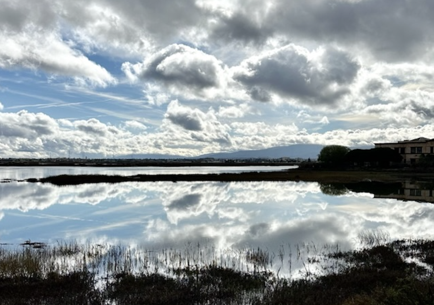 A reflective body of water mirrors a cloudy sky with blue patches, surrounded by dark vegetation and a building on the right, creating a serene landscape scene.