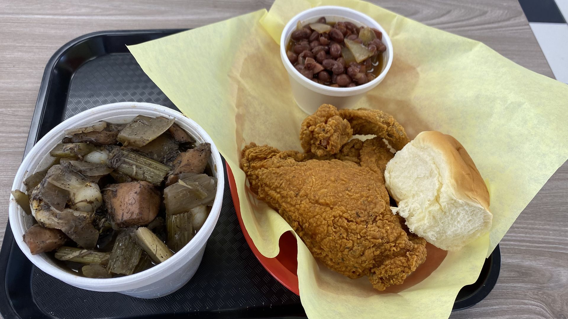 Fried chicken in a plastic basket, a Styrofoam cup of red beans and rice, and a Styrofoam bowl of gumbo on a black plastic tray.