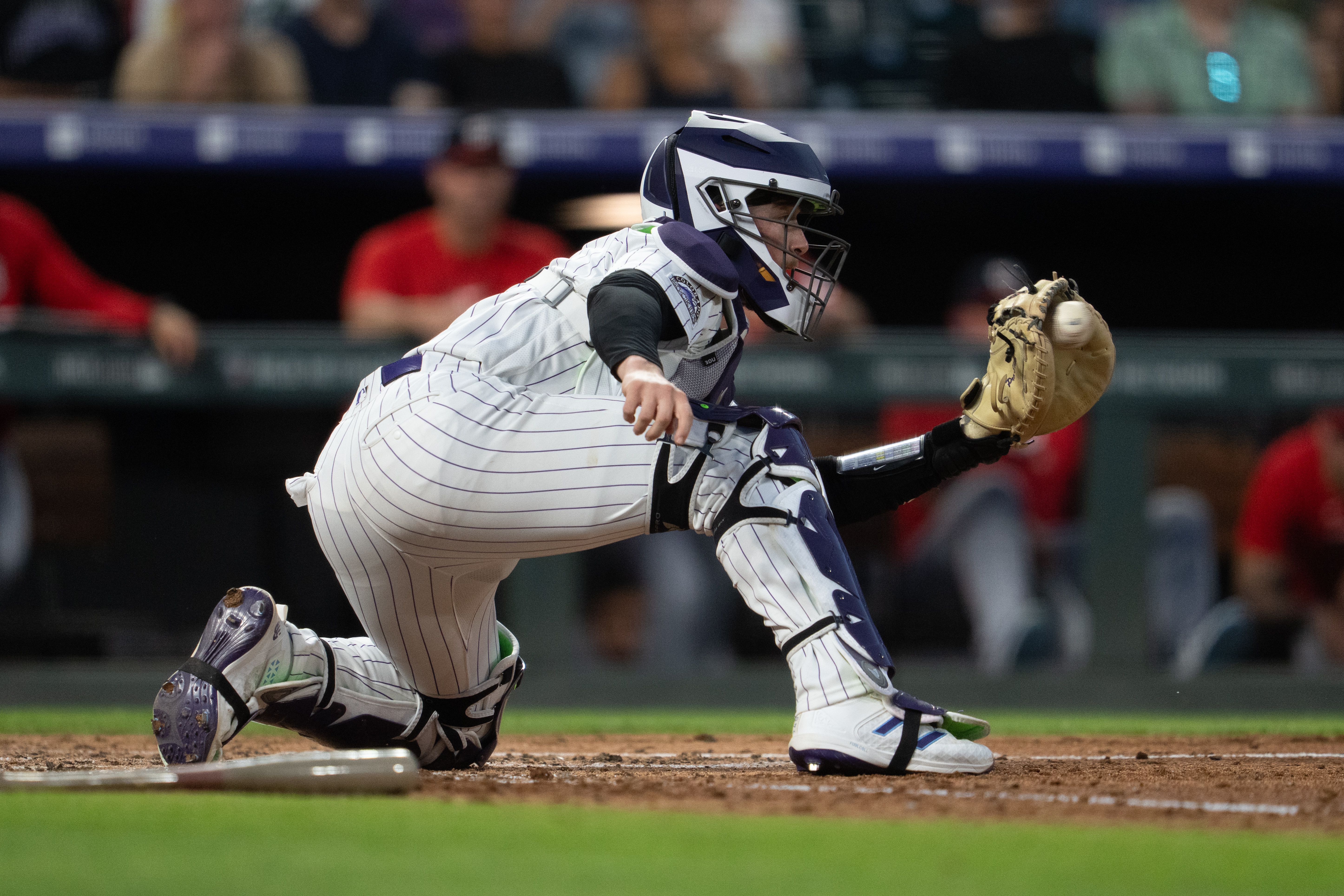 Baseball catcher in a white pinstriped uniform with purple accents, wearing helmet and mask, lunging toward home plate as the ball nears his glove; dirt, protective gear, and a blurred crowd in the background.