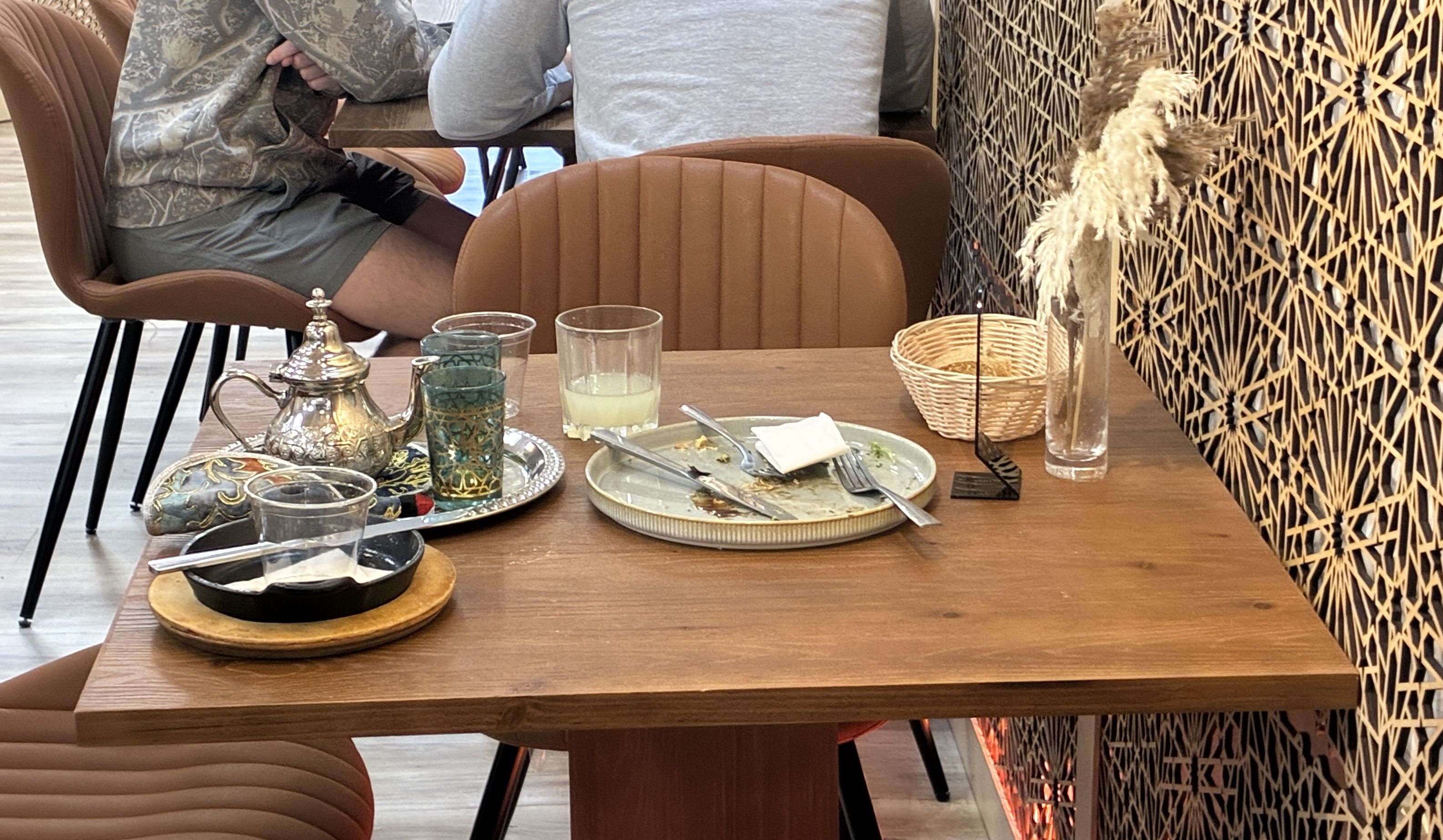 A restaurant table with a half-eaten meal, plates, forks, a silver teapot, glasses, a wicker basket and tall dried grasses, set against a bold geometric wall; two people sit with backs to the camera.