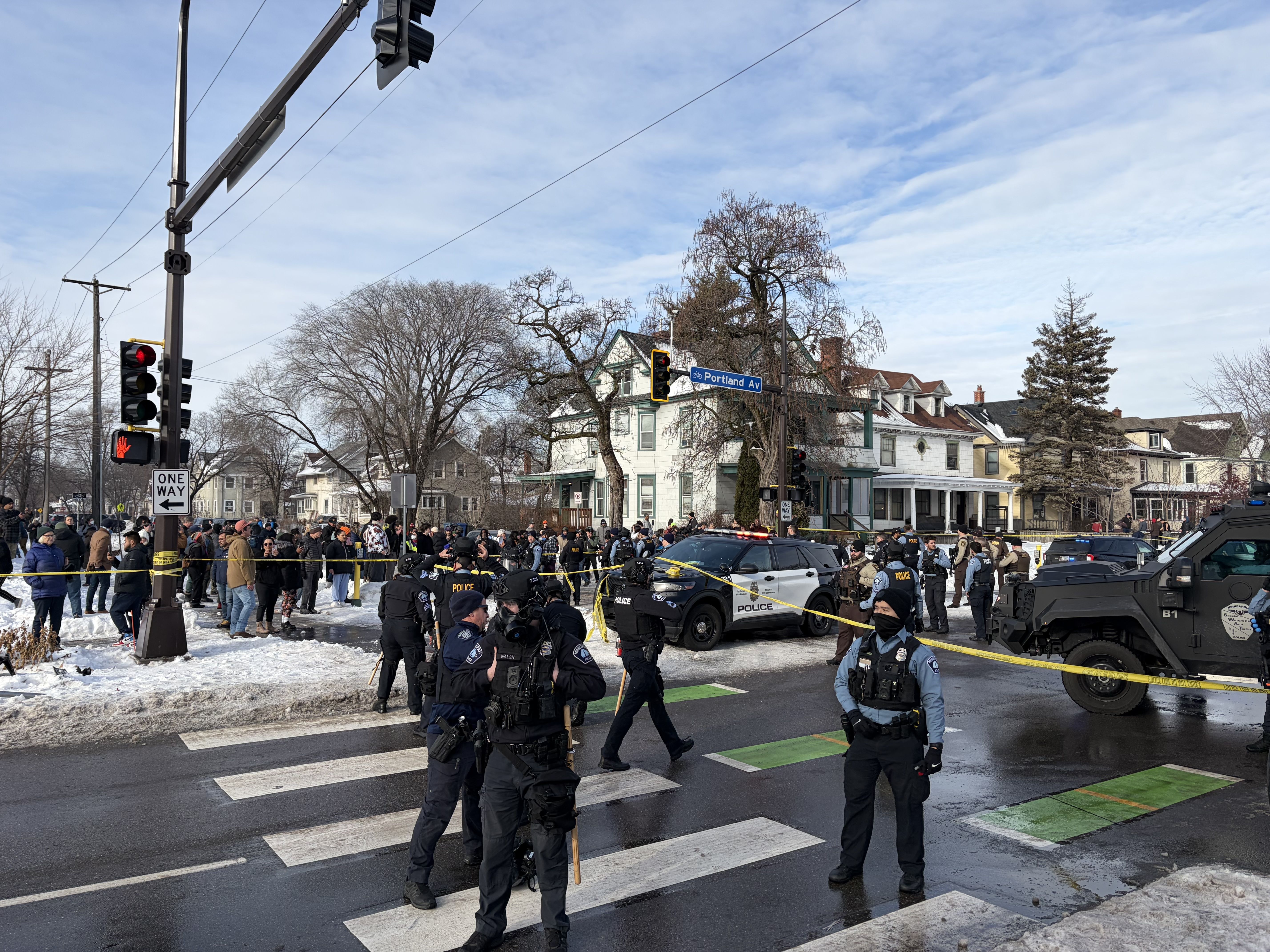 Police officers in gear block a snowy street at Portland Ave intersection with yellow caution tape, crowd gathered behind, police vehicles, and houses in the background under a partly cloudy sky.