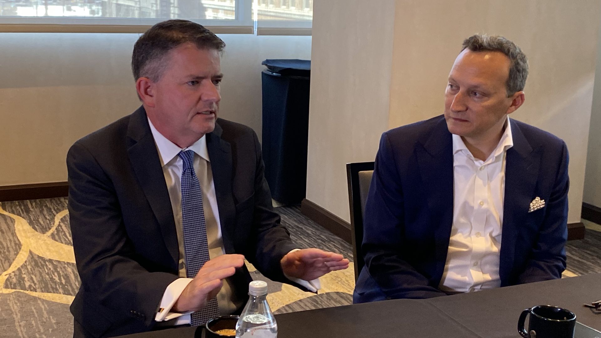Two men in suits sit at table while one of them talks and gestures with his hands. 