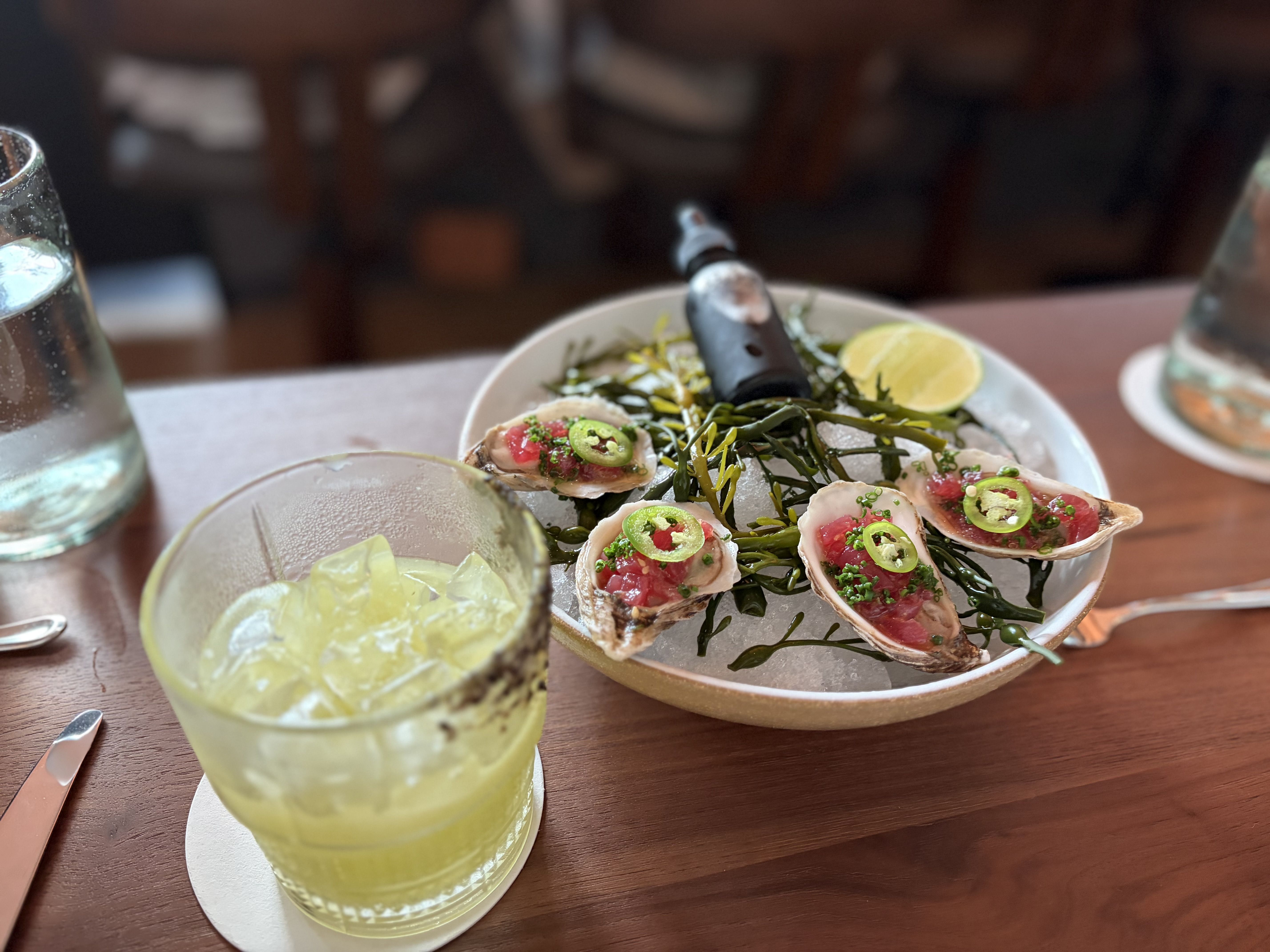 Plate of fresh oysters on a bed of seaweed, topped with chopped herbs and sliced green chiles; lime wedges on the plate. Two cold yellow beverages with ice in front, in a dining setting.