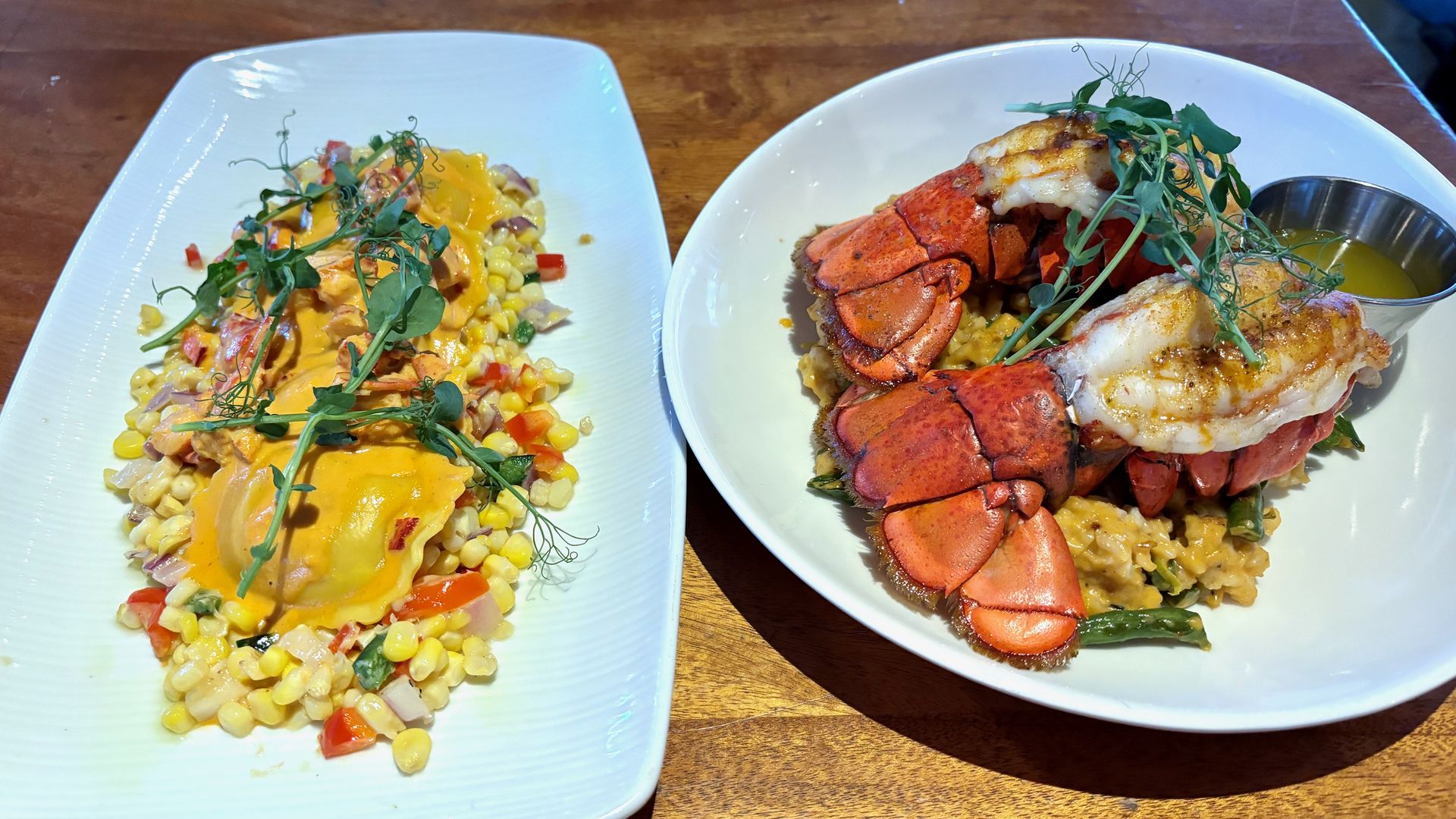 Two white plates, one with lobster ravioli over succotash and another with twin lobster tails over risotto