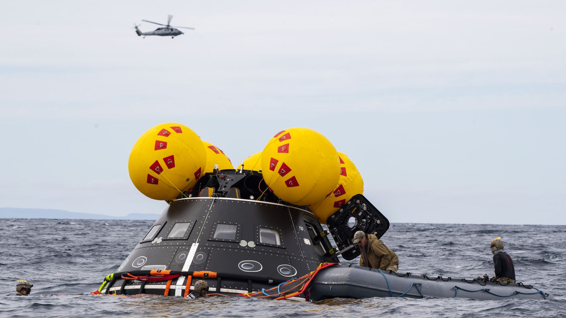 A dark rescue capsule partially submerged in the sea is tethered to an inflatable raft, with bright yellow balloons strapped to its top. Rescue crew in gear work nearby as a helicopter flies overhead.