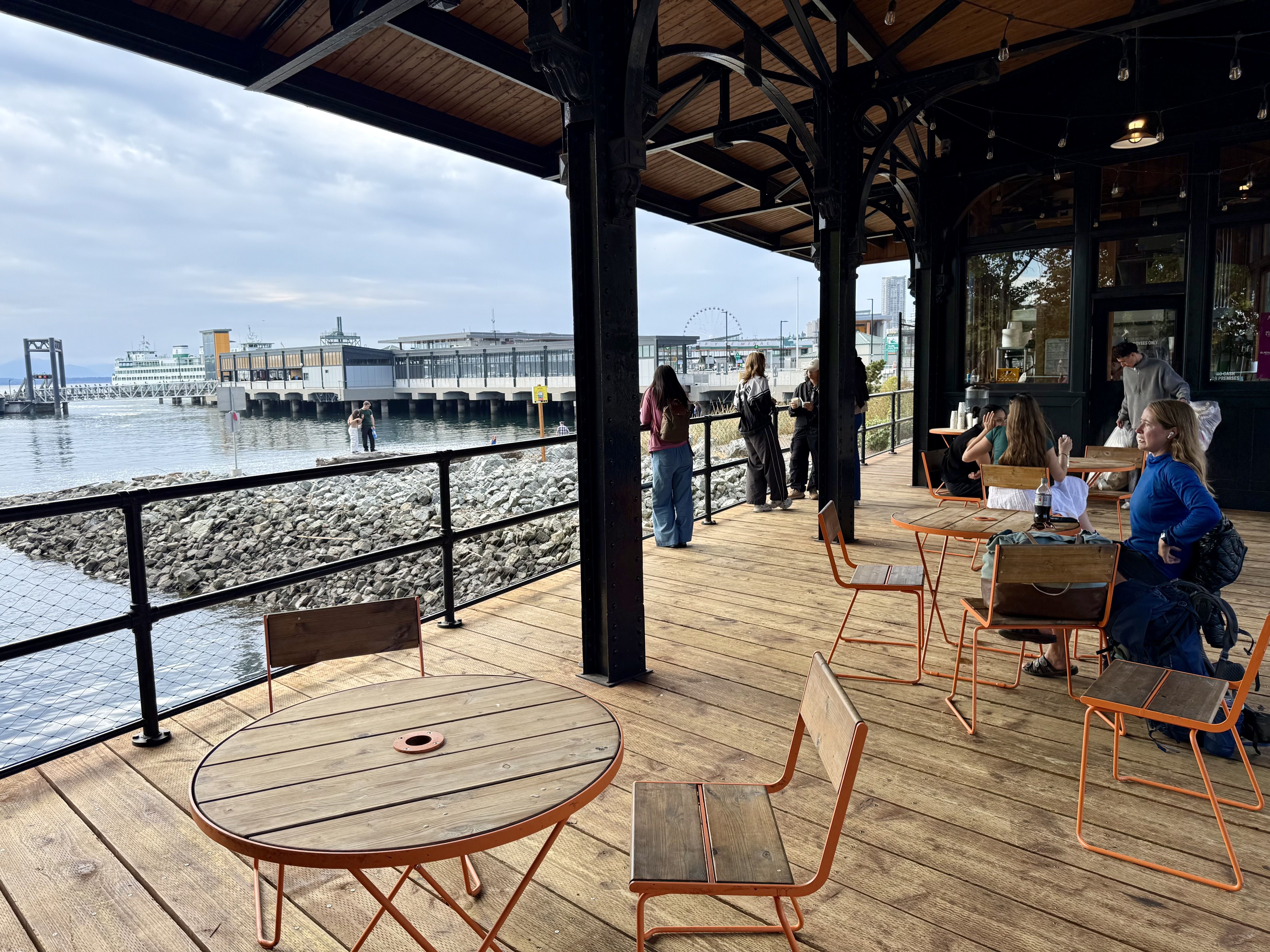 Outdoor wooden deck café with orange-framed tables and chairs, people chatting and looking out at rocky waterfront and distant ferry terminal under a cloudy sky.