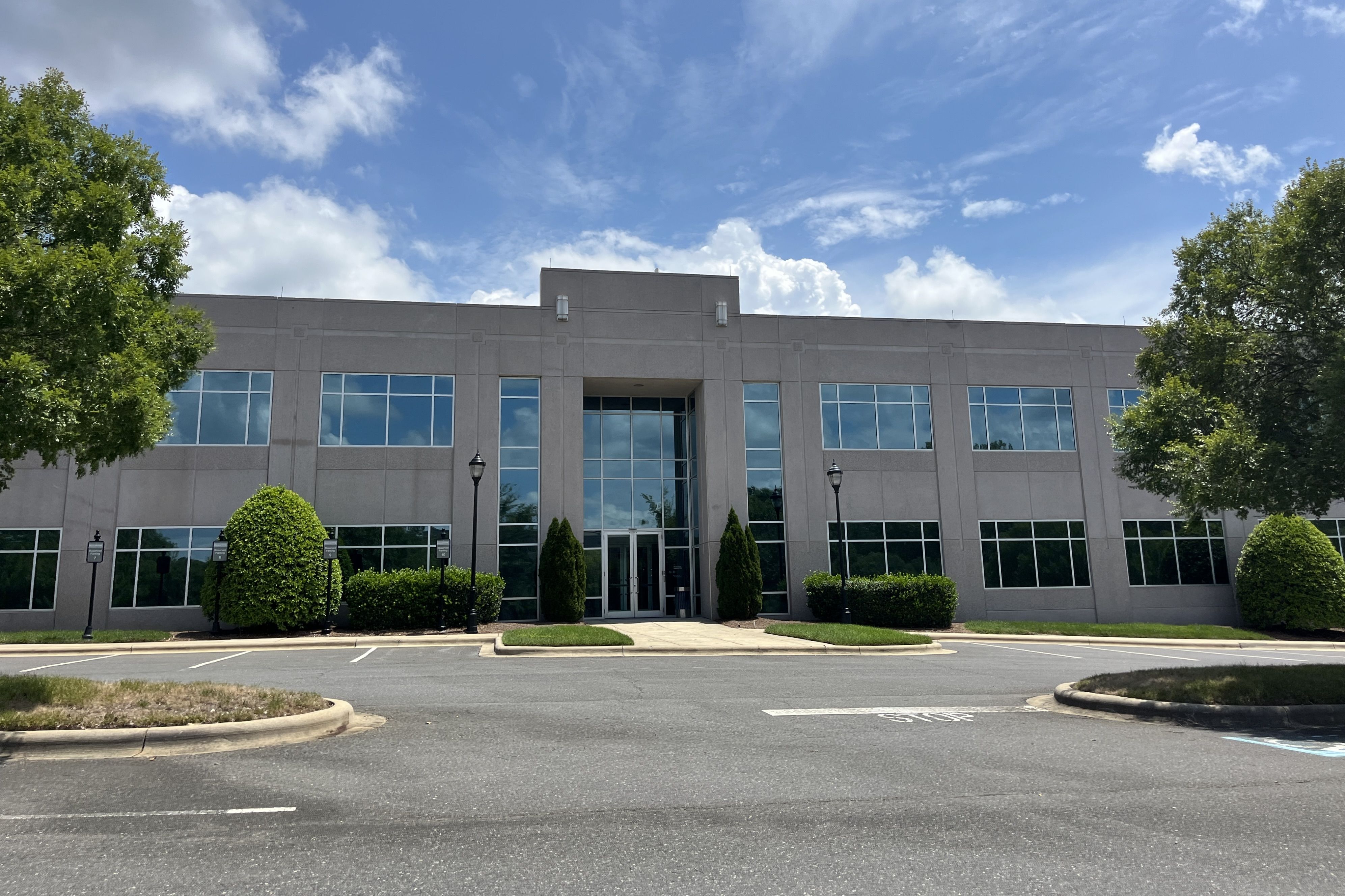 A modern gray office building with large glass windows and a central entrance, flanked by trees and shrubs, under a bright blue sky with white clouds.