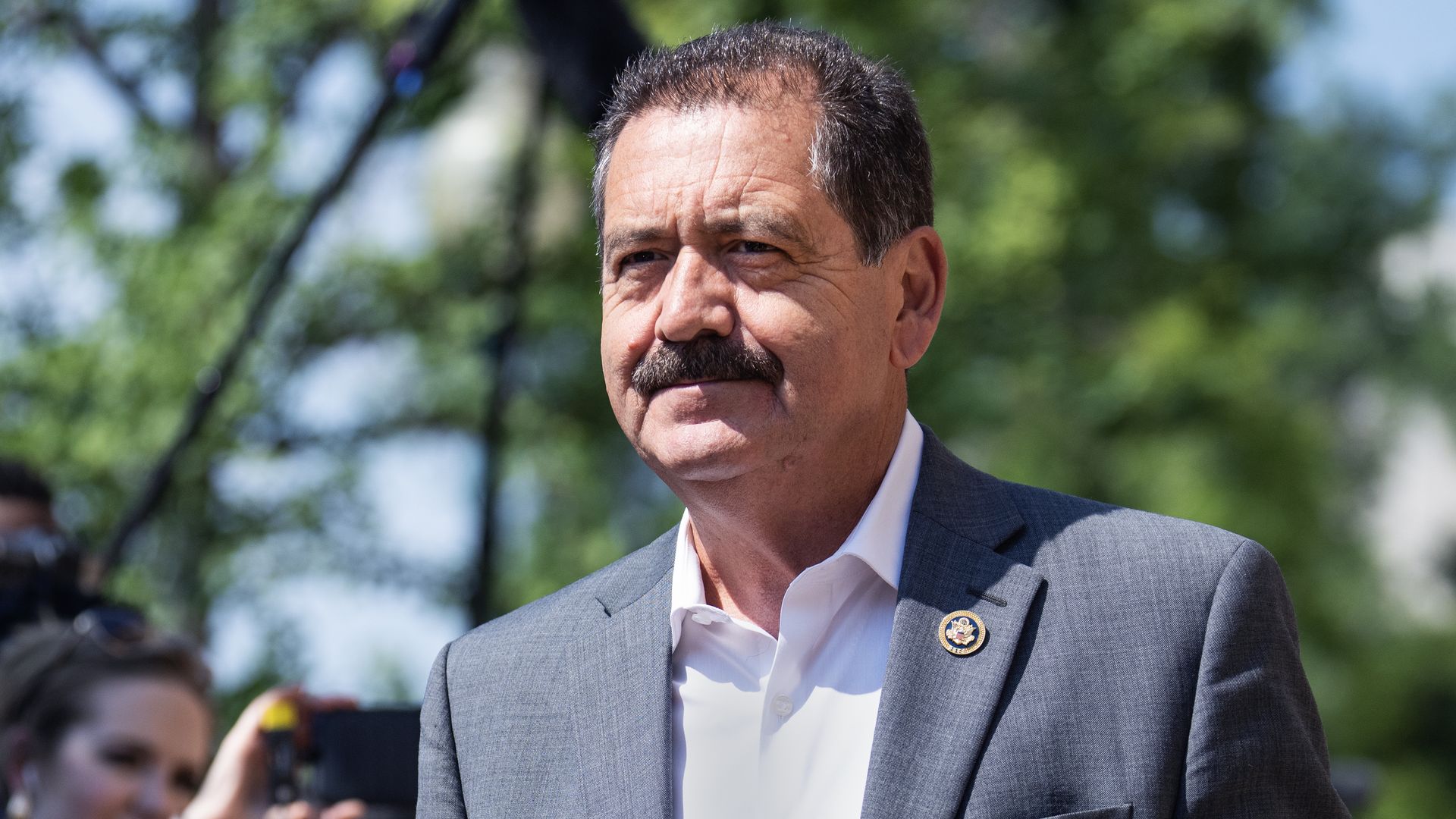 Rep. Chuy García, wearing a gray suit and walking past reporters outside on a tree-lined street.