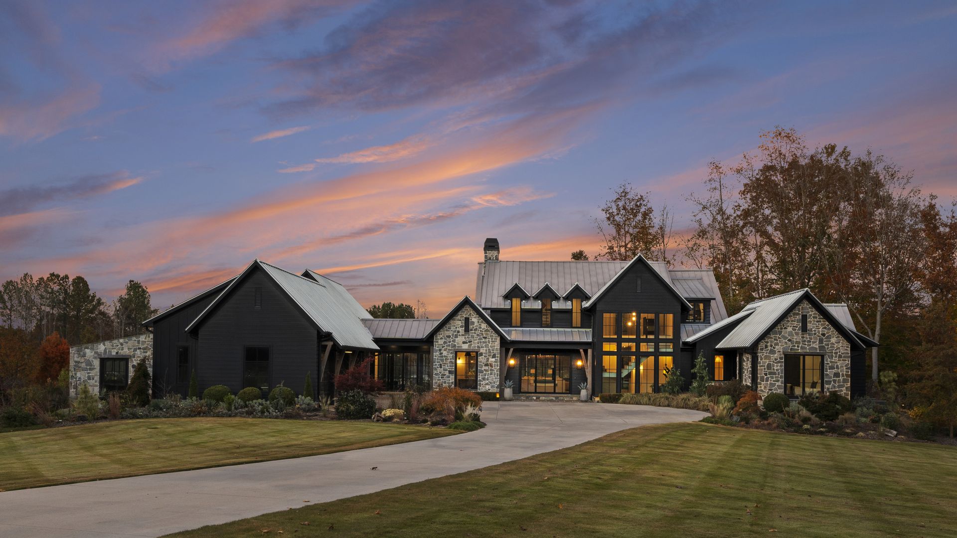 Luxurious modern house with black siding and stone accents sits along a curved driveway, warmly lit windows, autumn trees, and a pink-orange sunset in a spacious lawn setting.