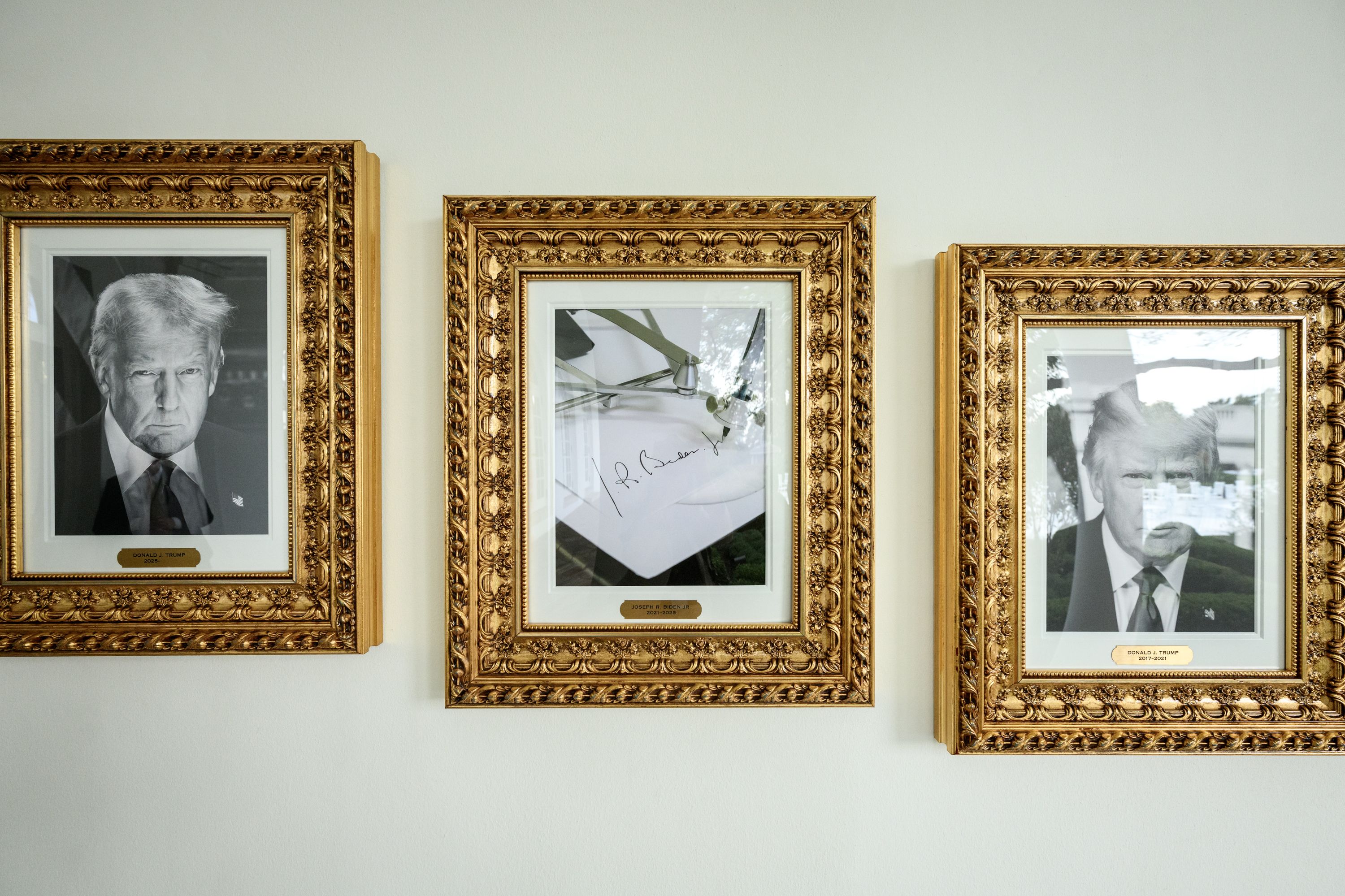 Three gold-framed images on a wall: left and right are black-and-white portraits of Donald J. Trump with plaques; center is a signed document with plaque for Joseph R. Biden Jr.