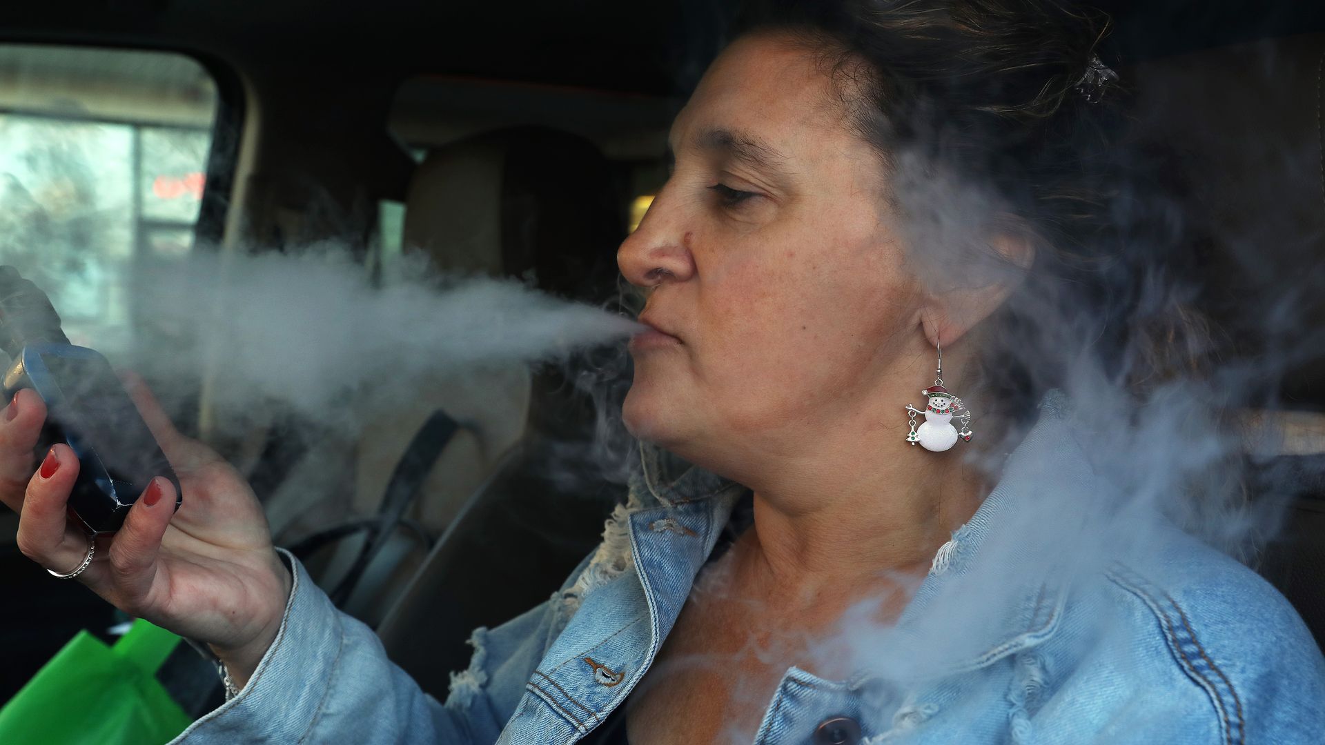 In this image, a woman wearing a jean jacket forms smoke while vaping with an e-cigarette.