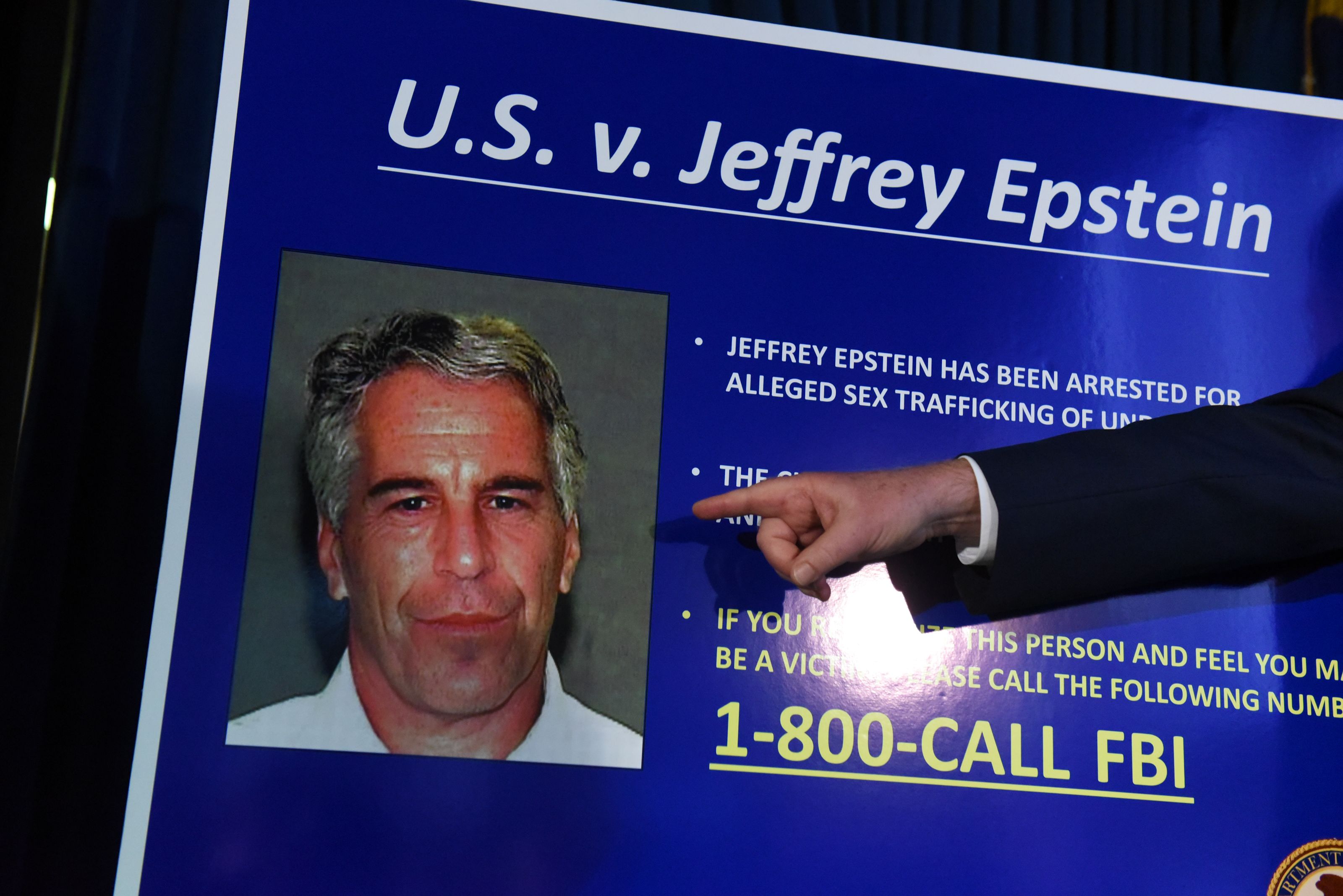 A sign titled "U.S. v. Jeffrey Epstein" with a photo of Epstein, text about his arrest for alleged sex trafficking, and a hand pointing to the FBI tip line 1-800-CALL FBI in yellow.