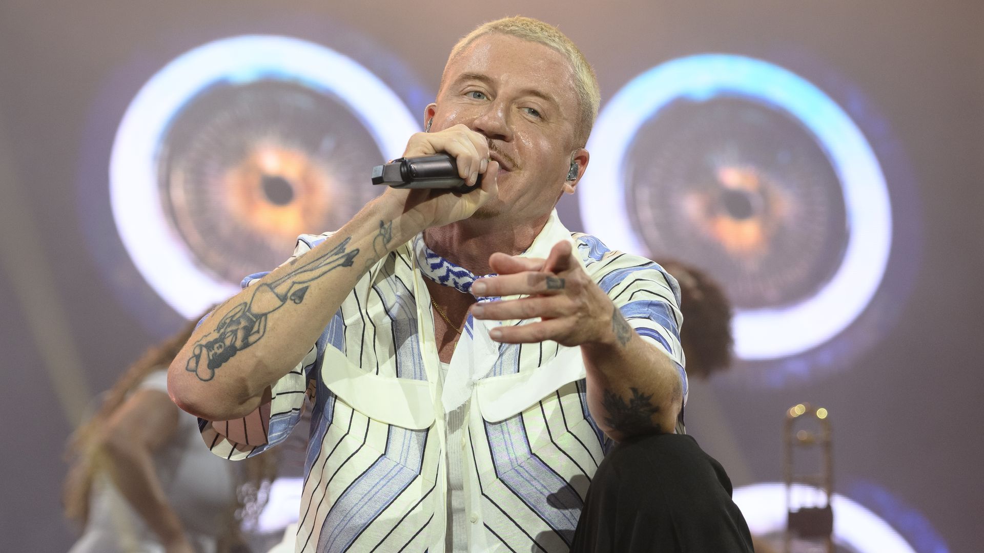 Macklemore performing holding a mic in his right hand  and pointing into the camera with his left hand. He's looking his left. 