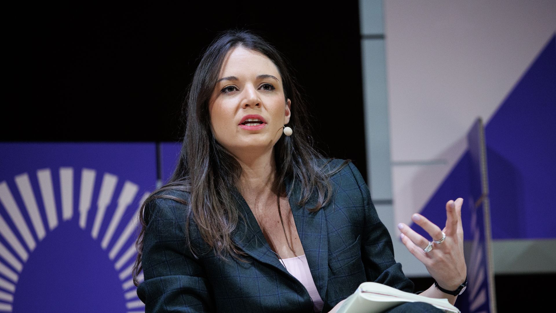 Irina Karamanos sits at an event with a small microphone near her mouth. she is holding her hand up and speaking. 
