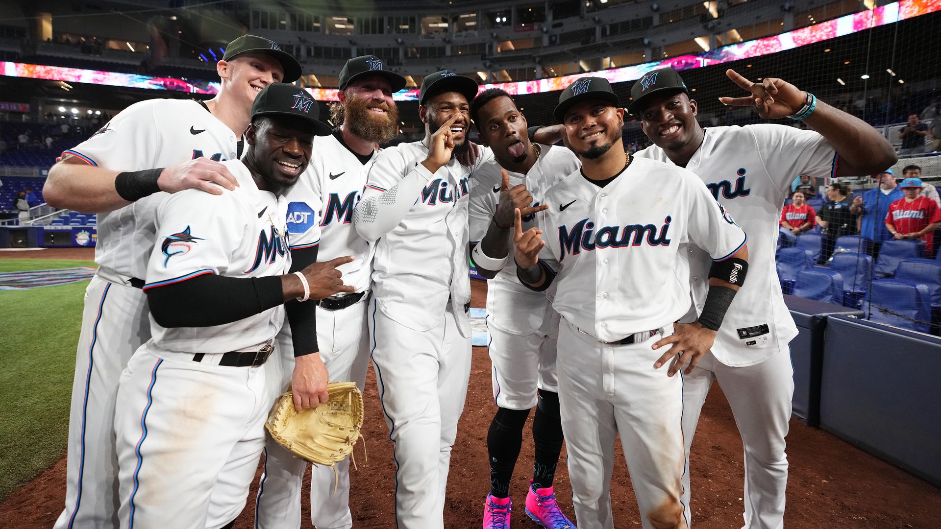 A group of baseball players in white uniforms gathers and smiles outside the dugout 