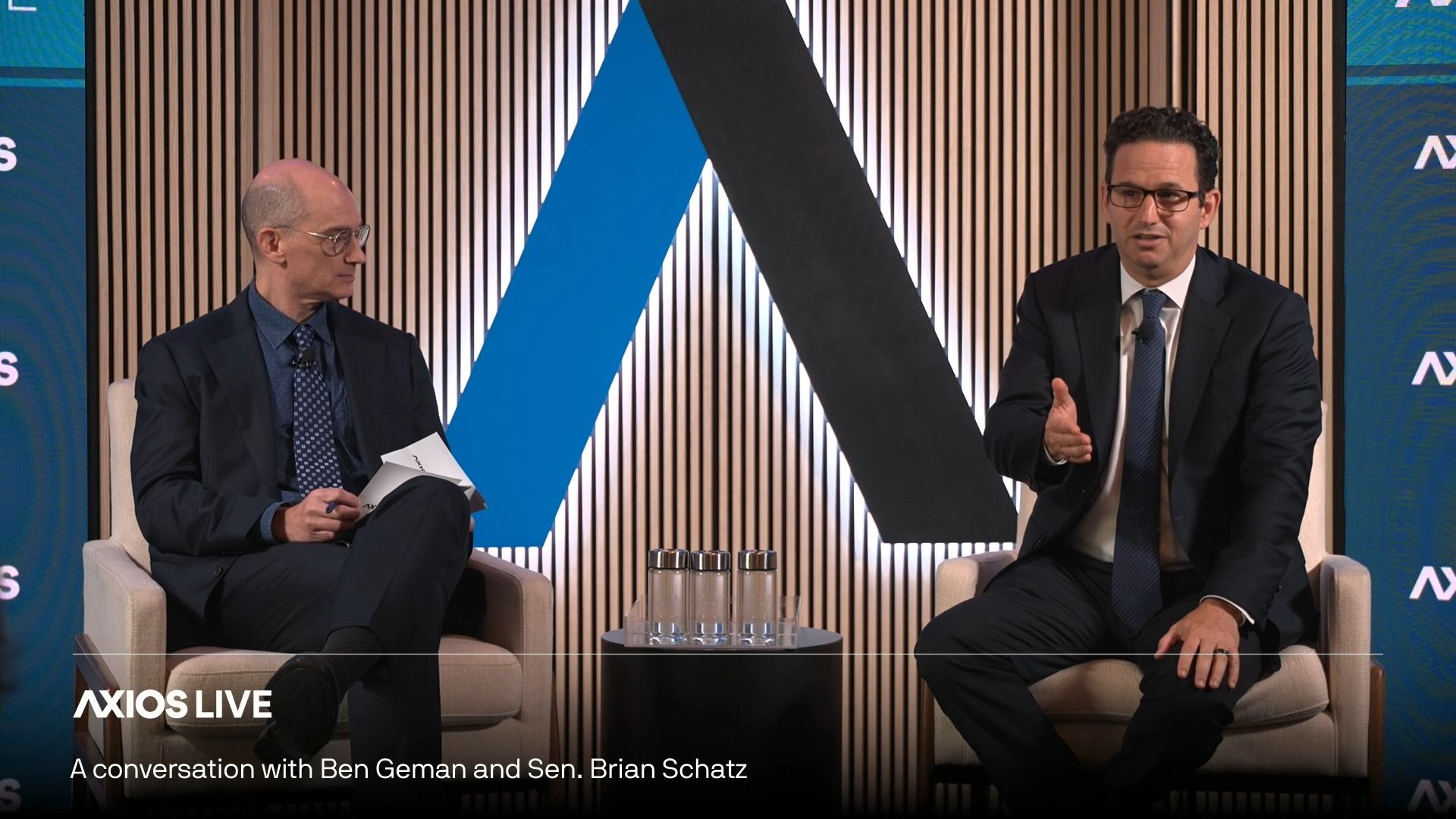 Two men in dark suits sit on cream chairs on a studio stage with vertical wood slats. A blue and black geometric backdrop sits between them; a glass table with water bottles is in front, Axios Live branding visible.