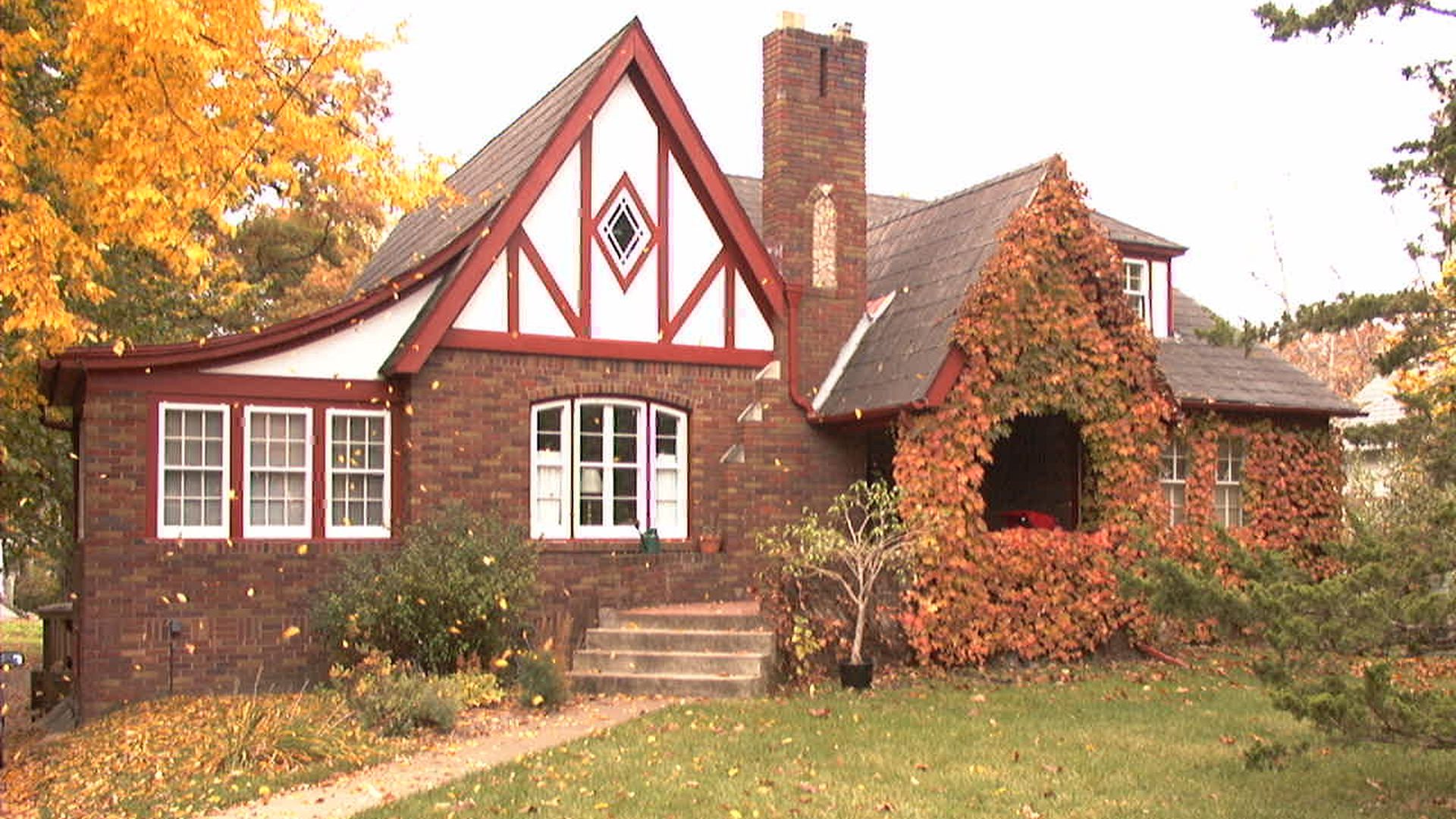 Autumn view of a brick Tudor-style house with white and red trim, a steep gable with a diamond window, a brick chimney, ivy-covered porch, orange trees, and fallen leaves on the lawn.