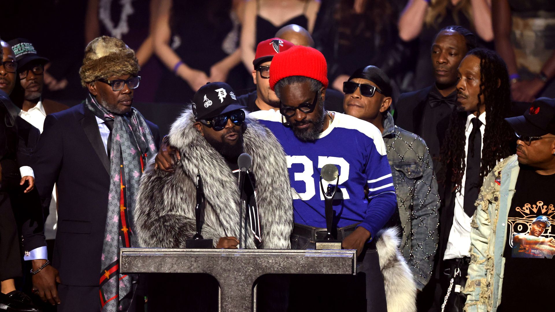 Group of men on stage at an award event, one in fur coat and sunglasses speaks at microphone, another in blue and white shirt with red beanie holds award, others stand behind them.