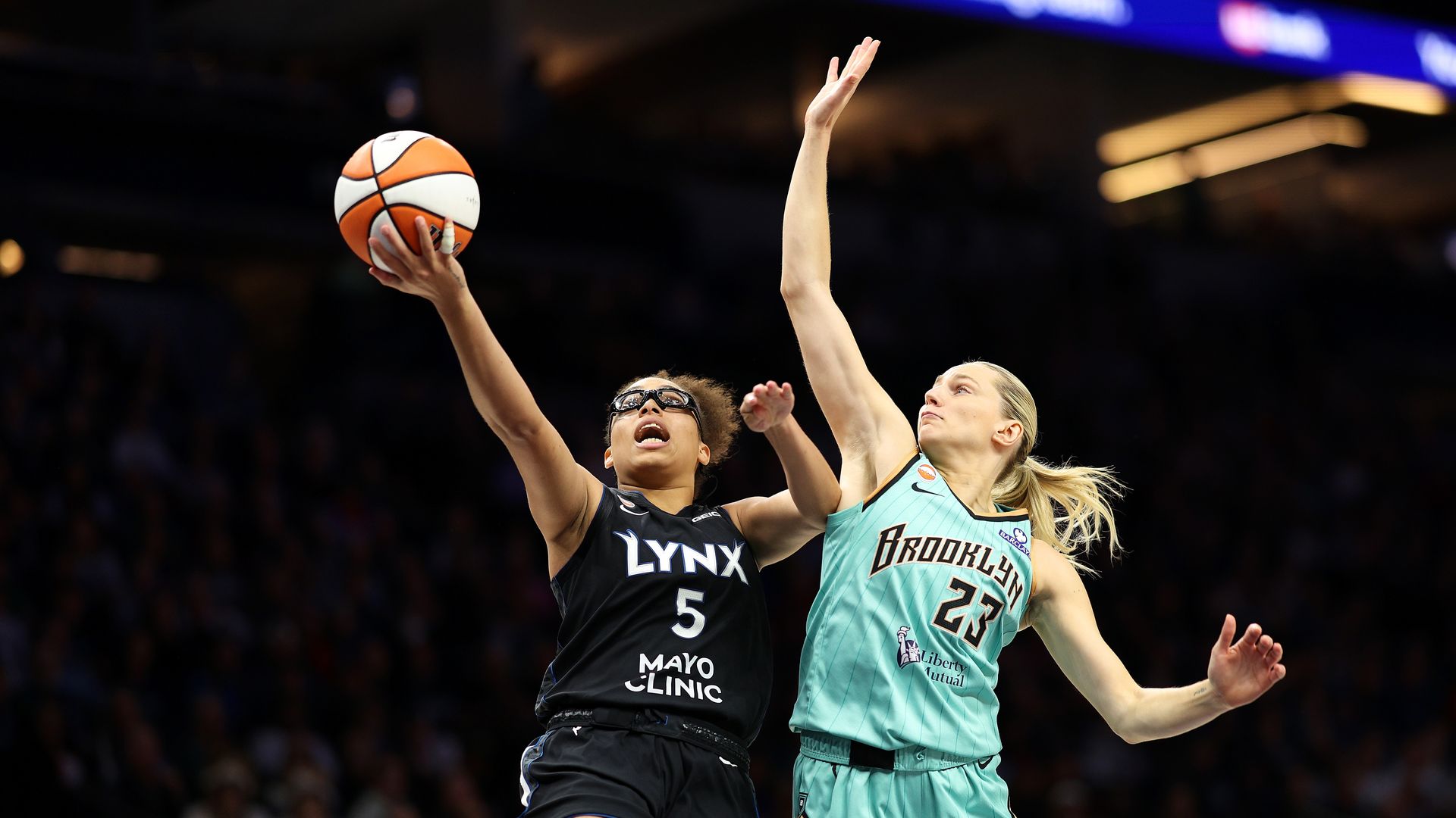Two female basketball players leap toward the ball in a game: one in a dark Lynx jersey (No. 5) reaches up, the other in teal Brooklyn jersey (No. 23) defends; blurred crowd in background.