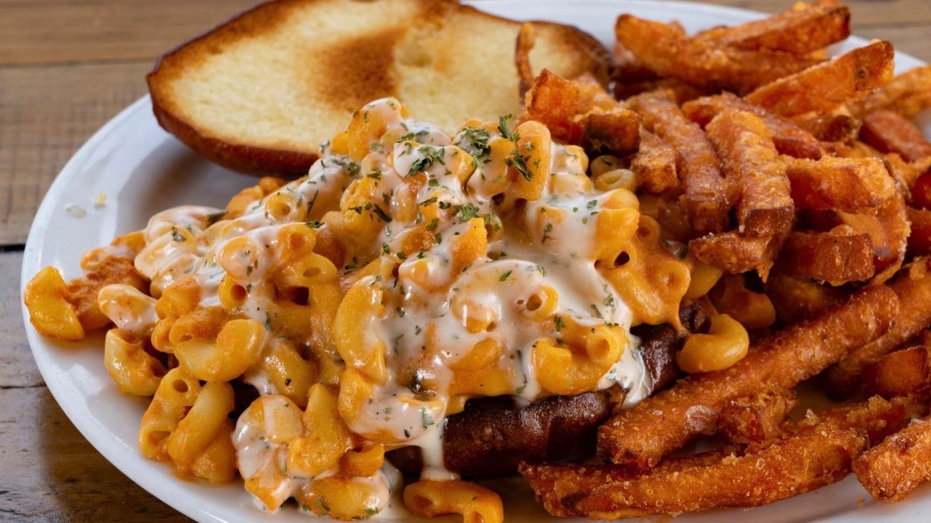 Plate of creamy macaroni and cheese with orange cheese sauce and herbs, next to crispy fries and a toasted bread slice on a white plate.
