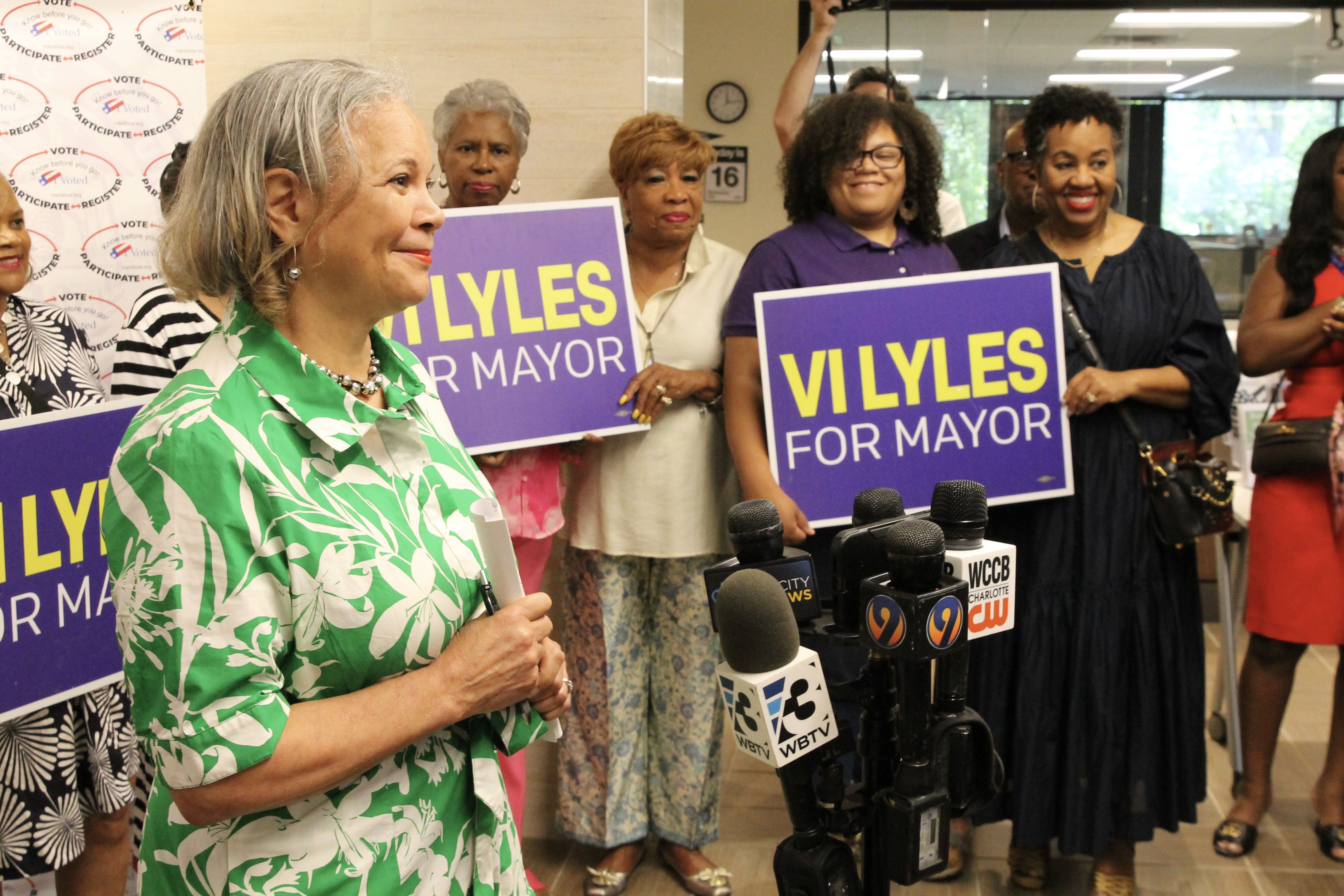 Woman in green and white floral dress speaking at a press event with supporters holding purple and yellow "VI LYLES FOR MAYOR" signs behind her, with microphones in front.