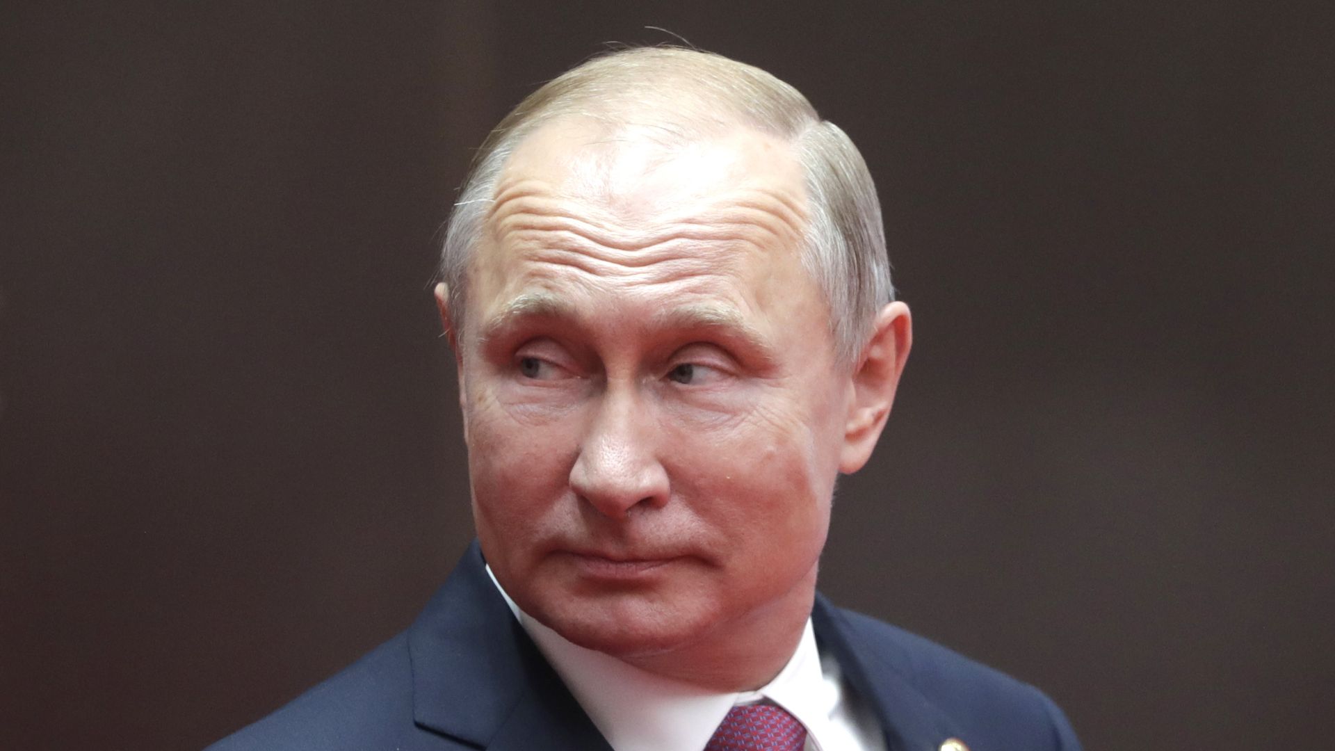 Vladimir Putin in a navy suit looks sideways before a dark background.