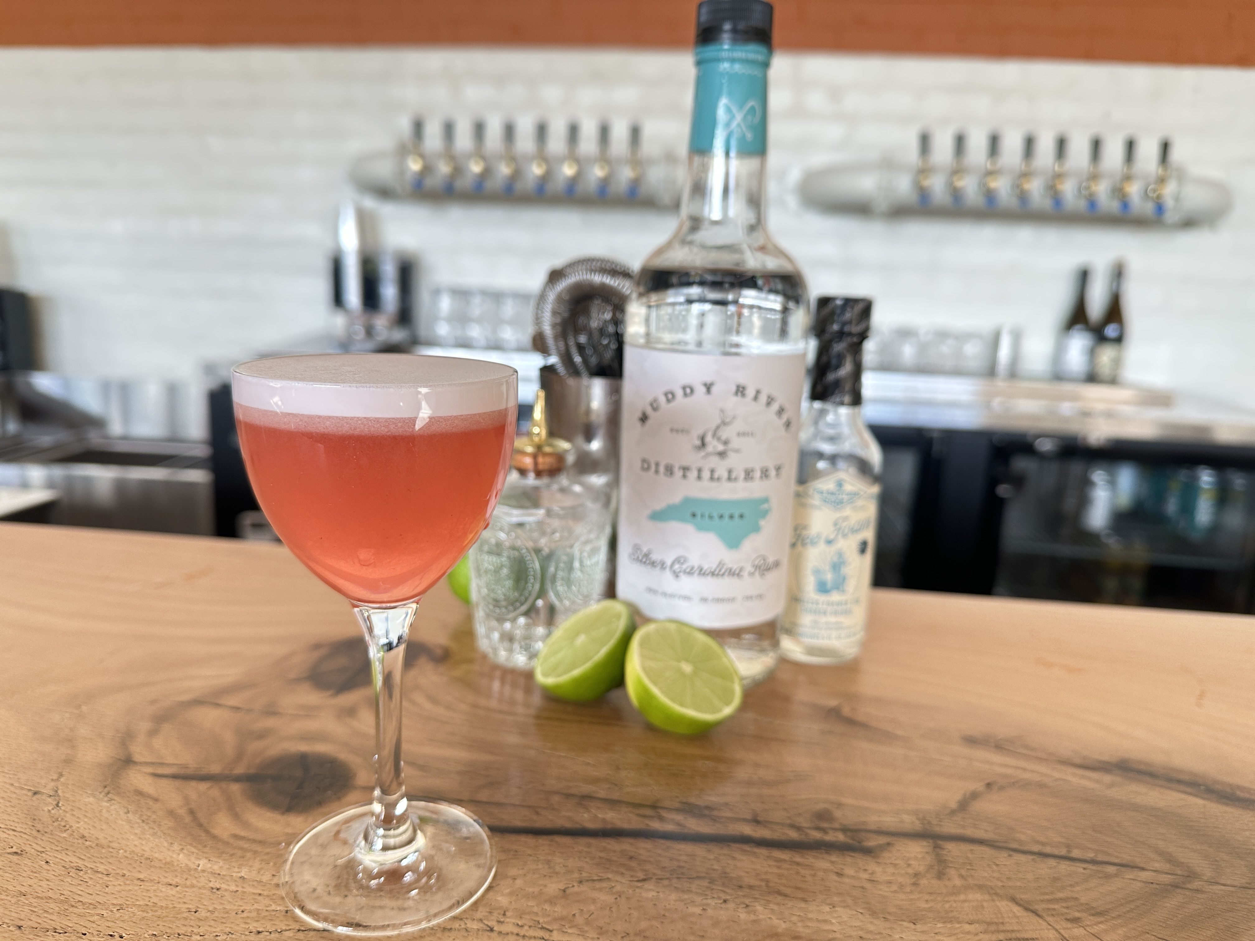 Pink cocktail in a stemmed glass sits on a wooden bar, with lime wedges nearby; blurred bar scene in the background shows a teal-labeled bottle and other bottles.and/or cafe is a new concept from former Lorem Ipsum owner that opens in Belmont neighborhood in the former Change Please cafe space inside Innovation Barn.