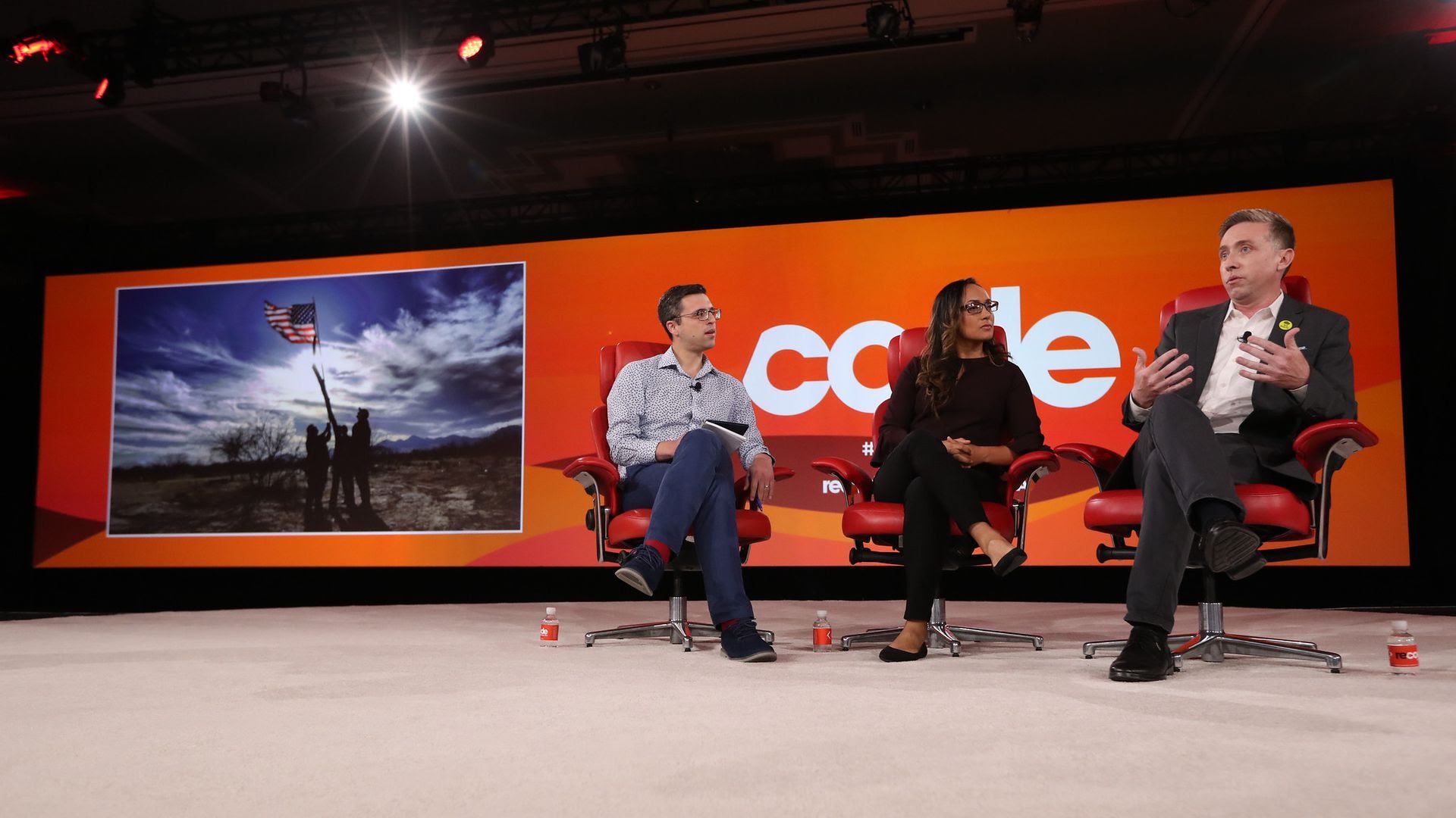Vox's Ezra Klein, RAICES' Erika Andiola and RAICES' Jonathan Ryan (from left to right) in office-like chairs with an orange banner reading "code" behind them.
