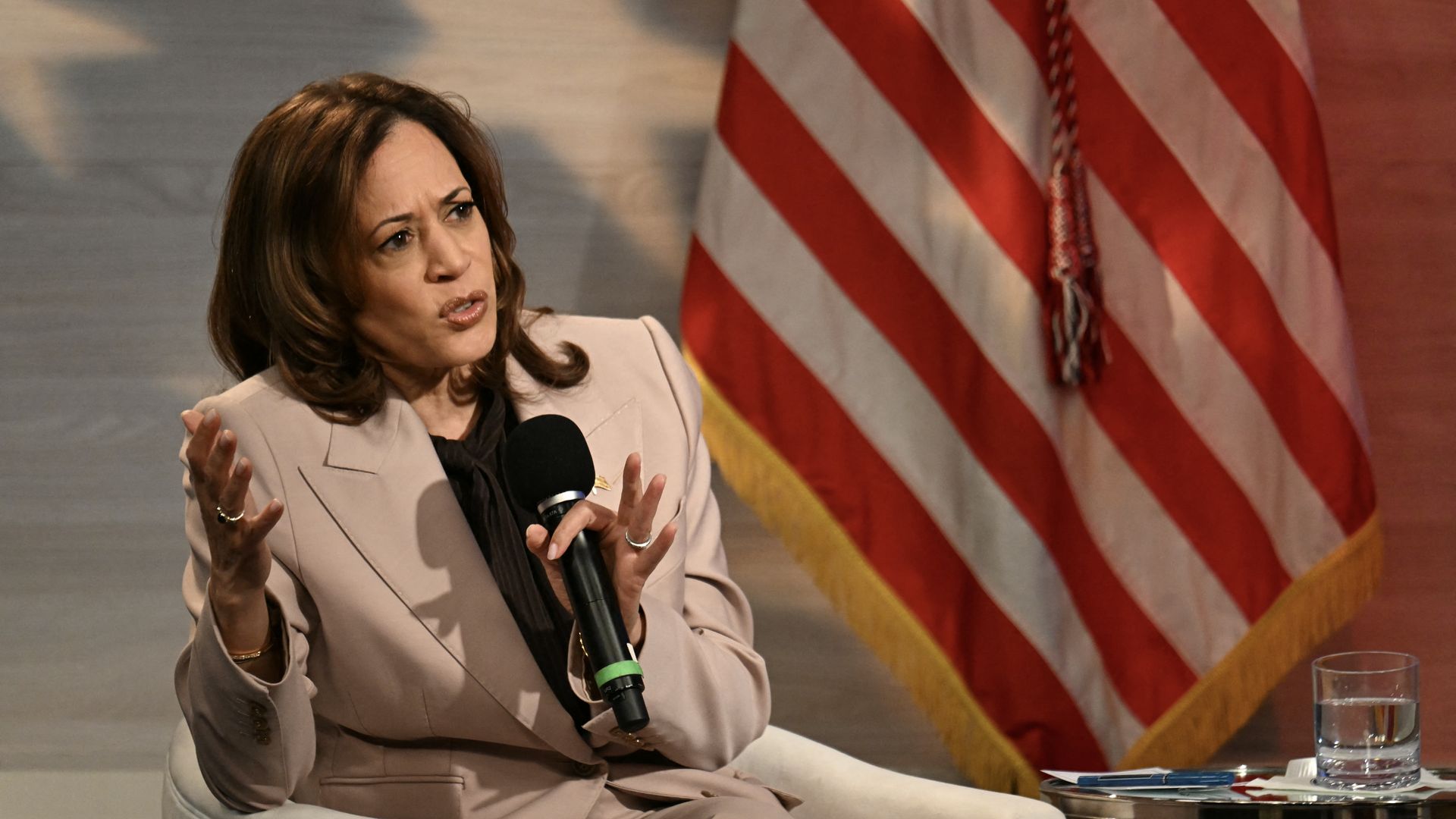 Kamala Harris, wearing a tan suit, holds a microphone. She is seated with an American flag behind her. 