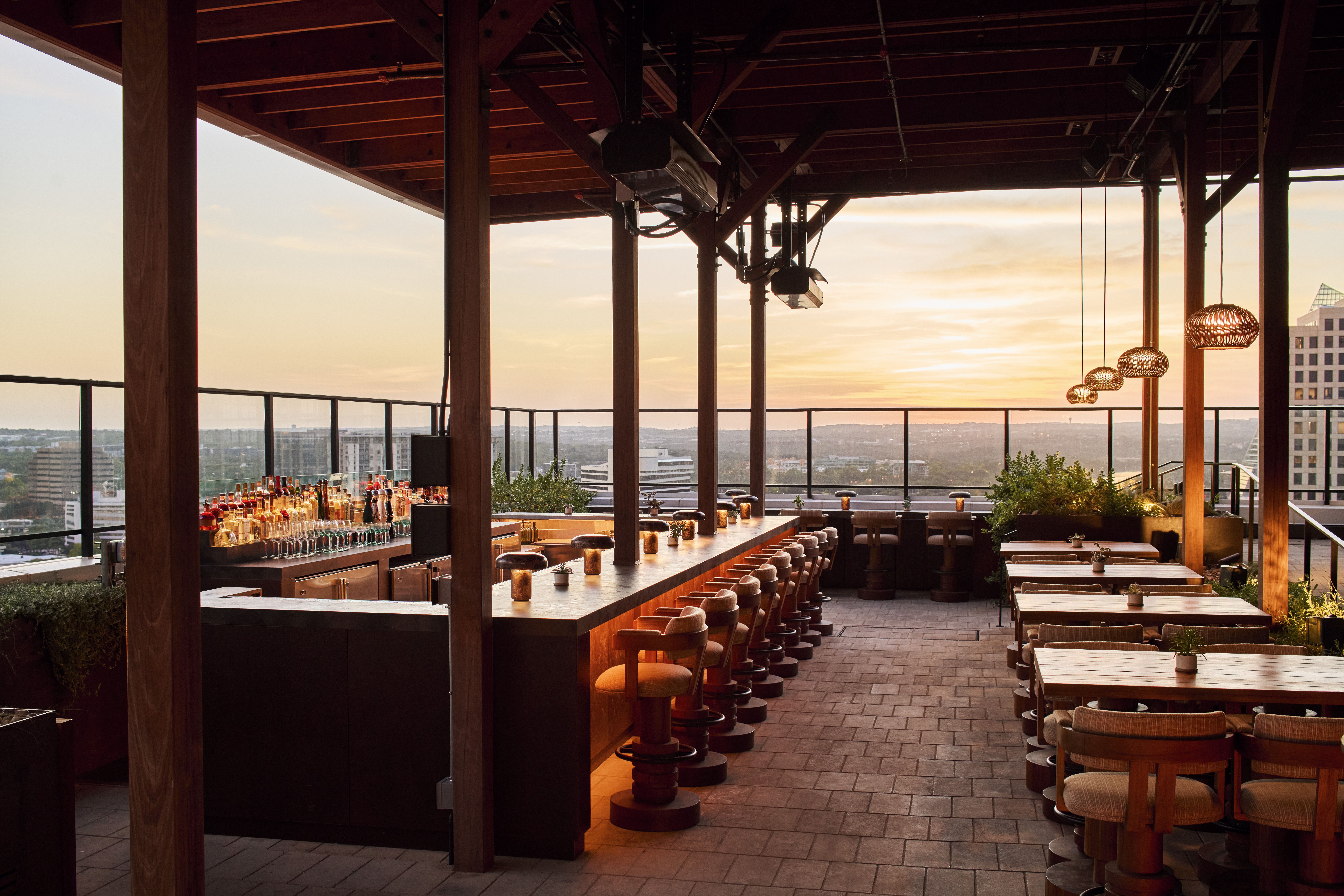 Sunset rooftop bar with a long wooden counter and stools, glass railing, and hanging wicker lamps. Bottles line the bar as a warm orange sky and distant city skyline glow.