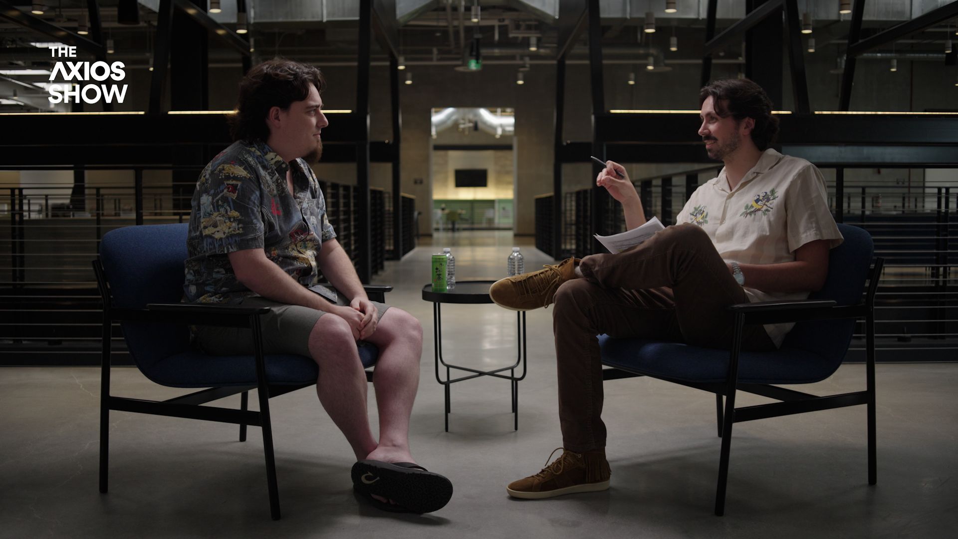 Two men sit in blue chairs facing each other in a modern, industrial space. A small round table between them holds water bottles; one gestures with a pen while the other listens. The "The Axios Show" logo is in the top-left.