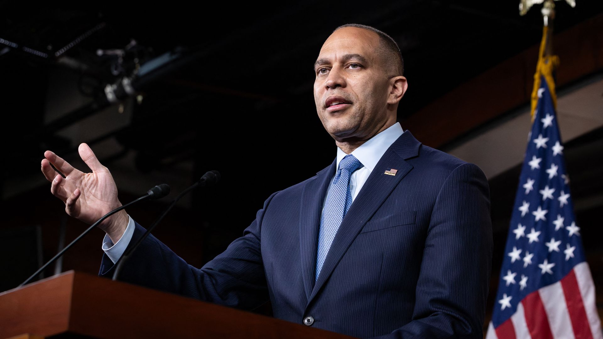 House Minority Leader Hakeem Jeffries, wearing a blue suit, stands at a podium in front of an American flag.