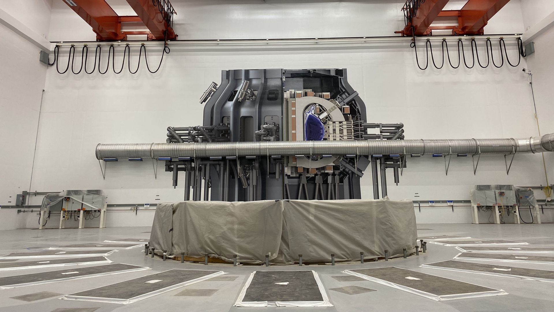 A photo of Tokamak Hall, a giant, mostly white-walled building at Commonwealth Fusion Systems' headquarters in Devens, Mass., where the company is building its next reactor. A series of gray stickers on the far wall depict roughly where the reactor will be and how it will look.