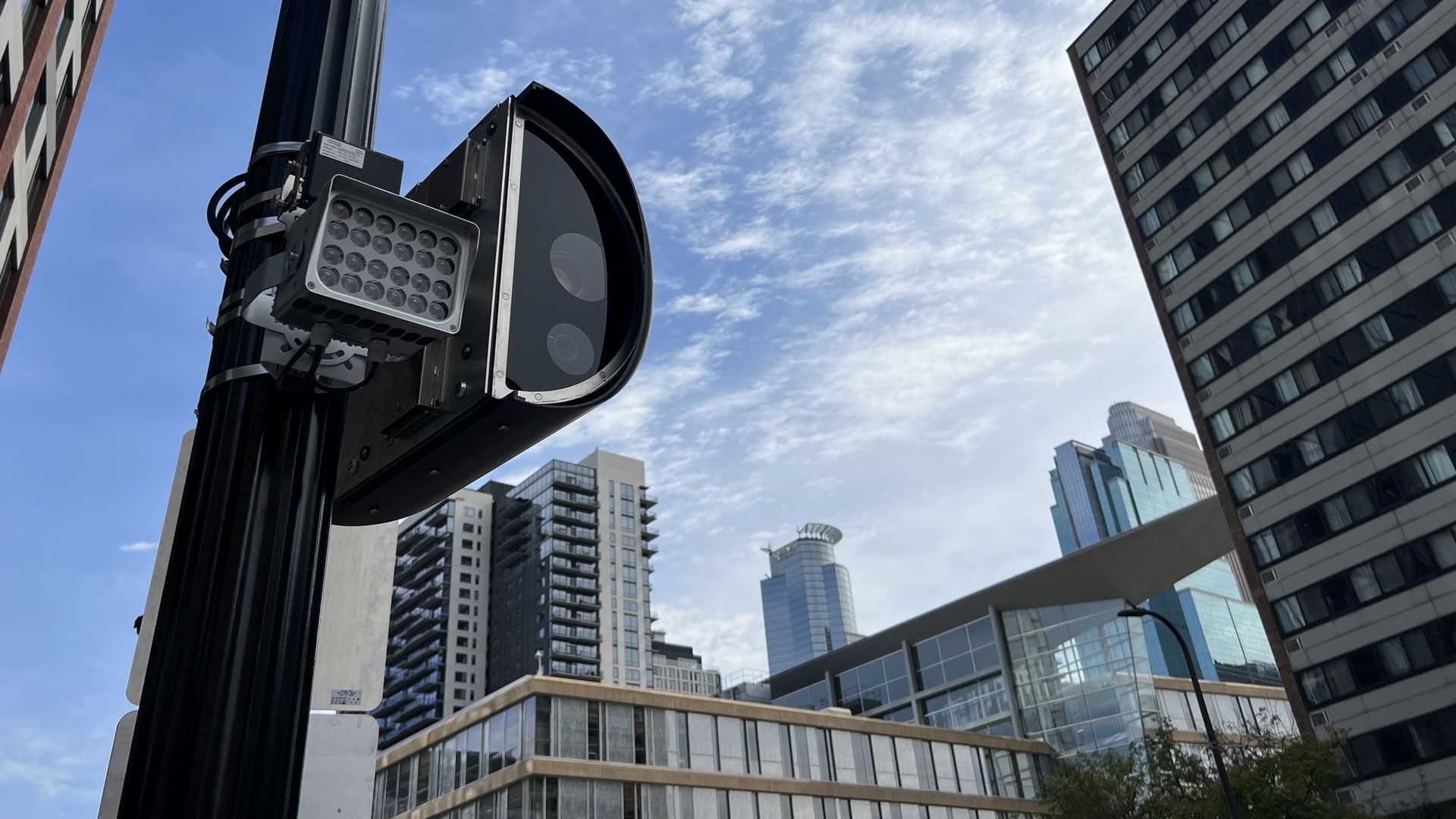 Close-up of a black traffic camera on a pole against a blue sky with clouds, surrounded by tall urban buildings in downtown Minneapolis with glass windows and varied architecture.