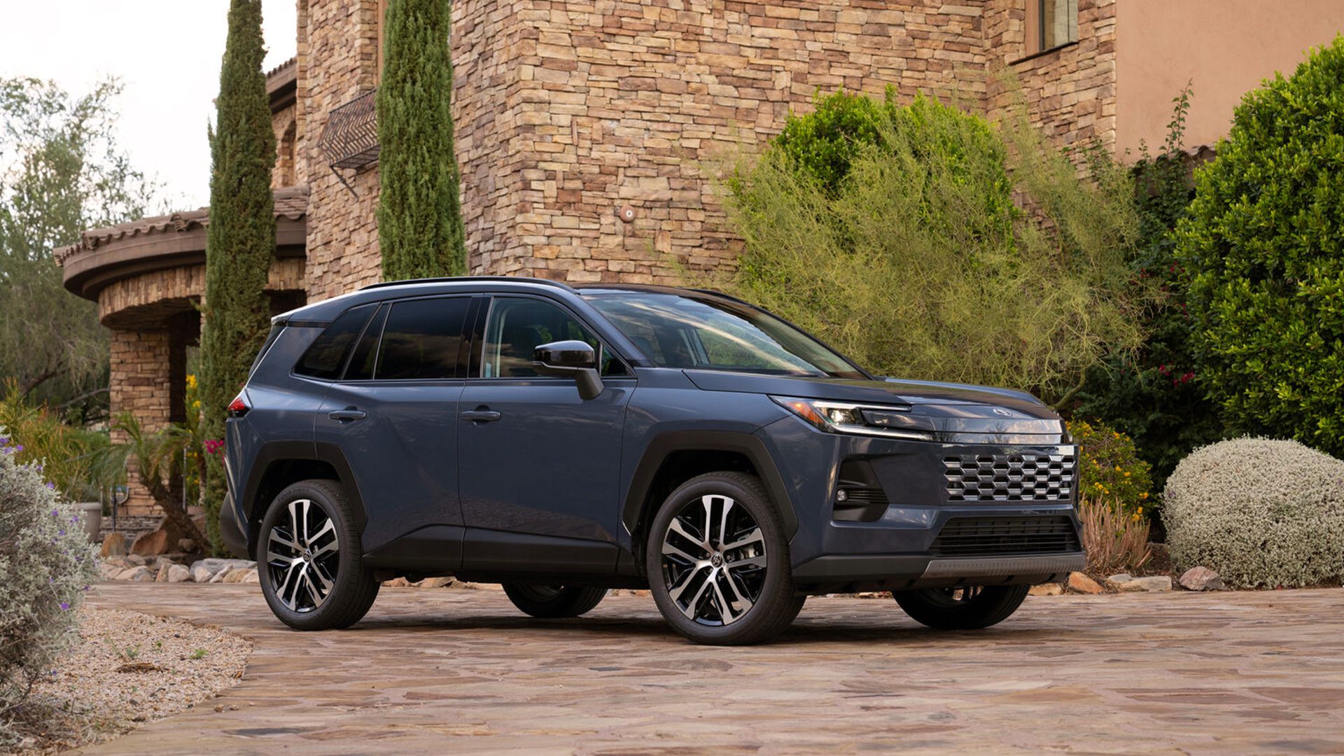 Blue Toyota Rav 4 SUV parked on a stone driveway in front of a tan stone house with tall cypress trees and desert landscaping.
