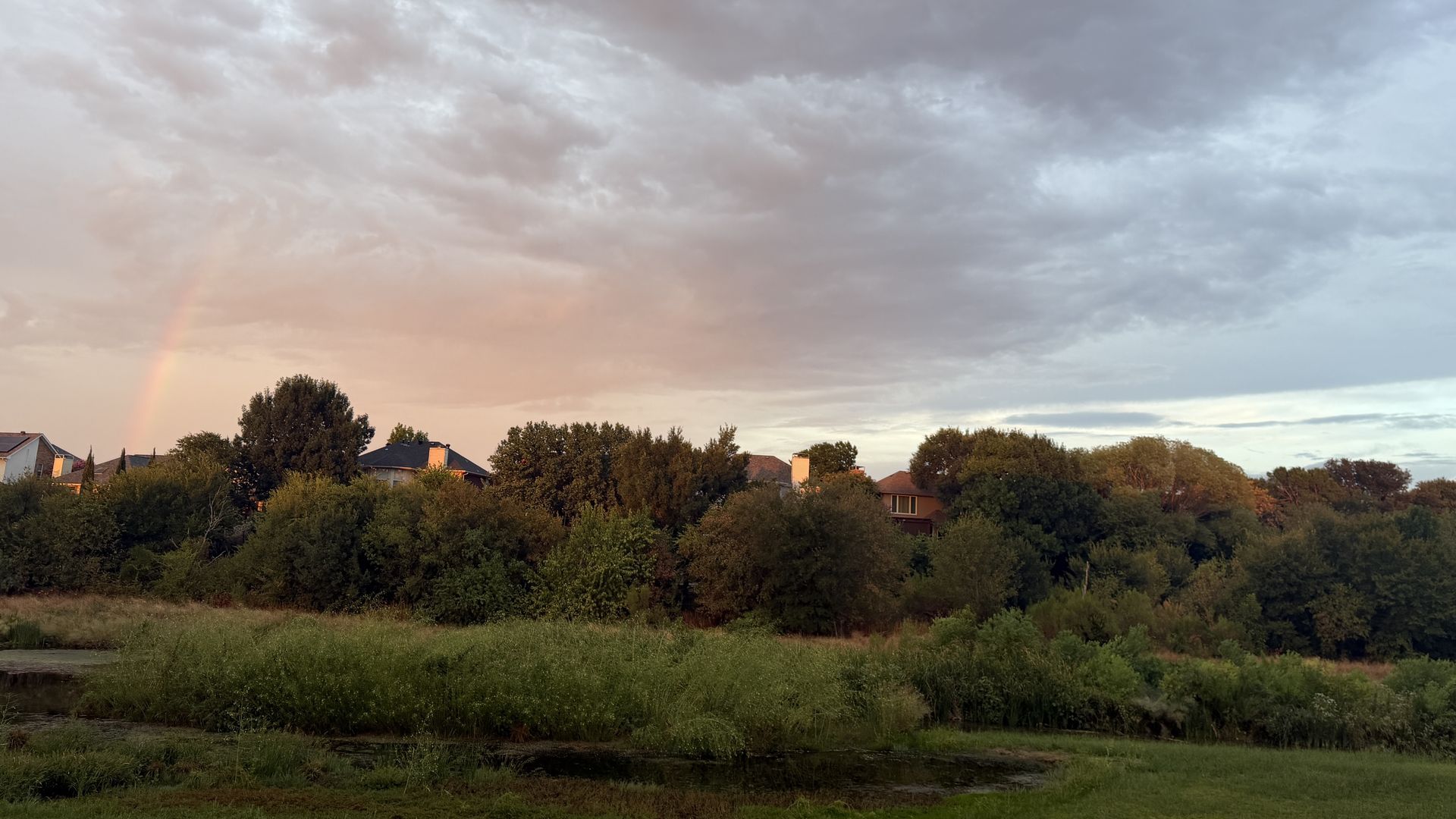 Evening view of a suburban skyline with trees and houses along the horizon; a faint rainbow appears on the left under a cloudy sky, over a grassy foreground.