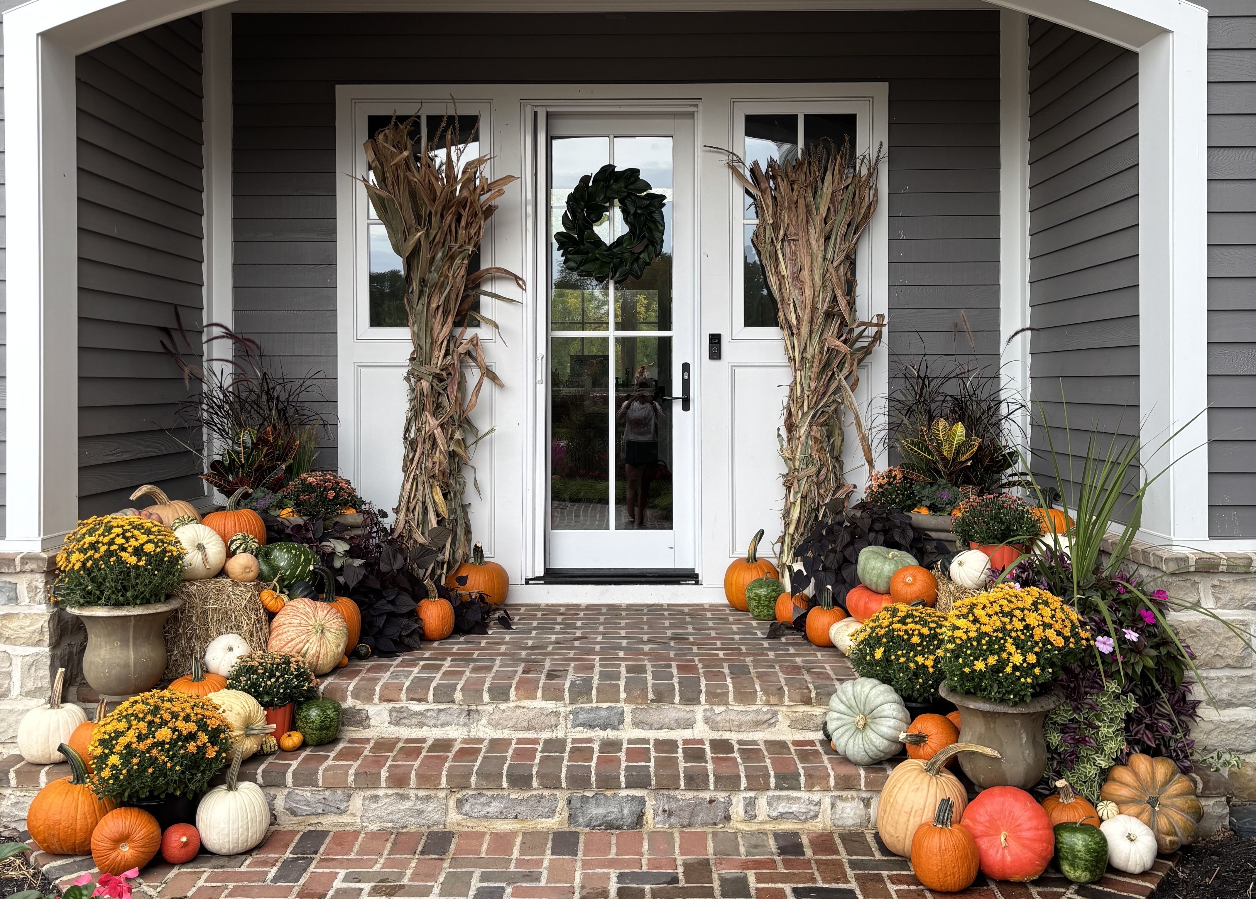House entrance decorated with orange, white, and green pumpkins, yellow flowers, cornstalks, and a green wreath on a glass door, creating a festive fall display.