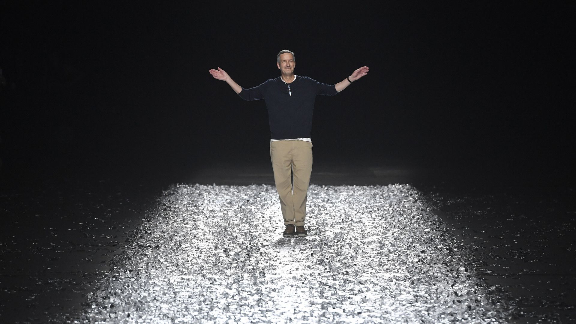 man standing on a catwalk wearing dark blue sweater and light colored pants waving with both hands to crowd