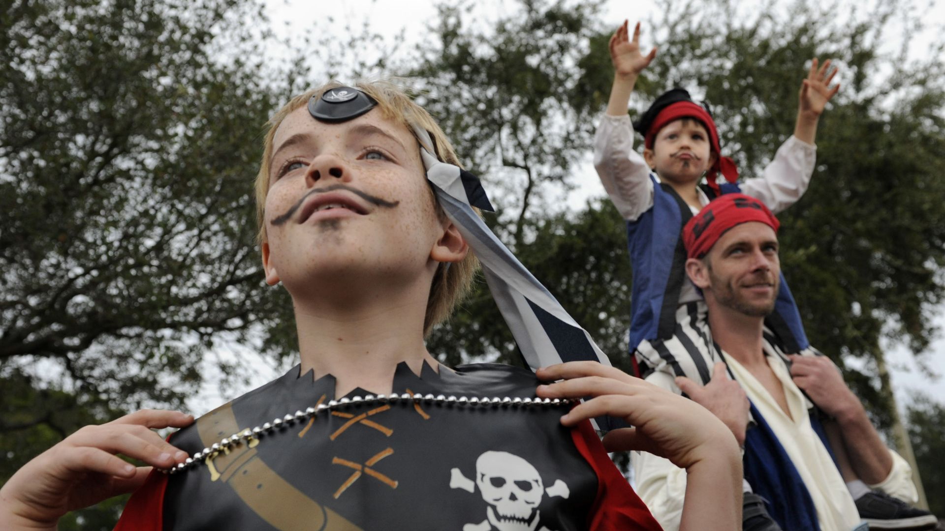 Boy in foreground wearing a black pirate costume with a skull emblem and a striped headscarf, with mustache face paint. Behind him, a man in pirate clothing carries a smaller child dressed as a pirate with arms raised outdoors.