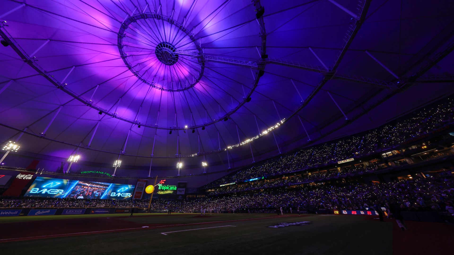 Interior of Tropicana Field, a domed baseball stadium bathed in purple light. A large circular roof ring and radial supports dominate the ceiling, with bright LED boards and a full crowd around the outfield.