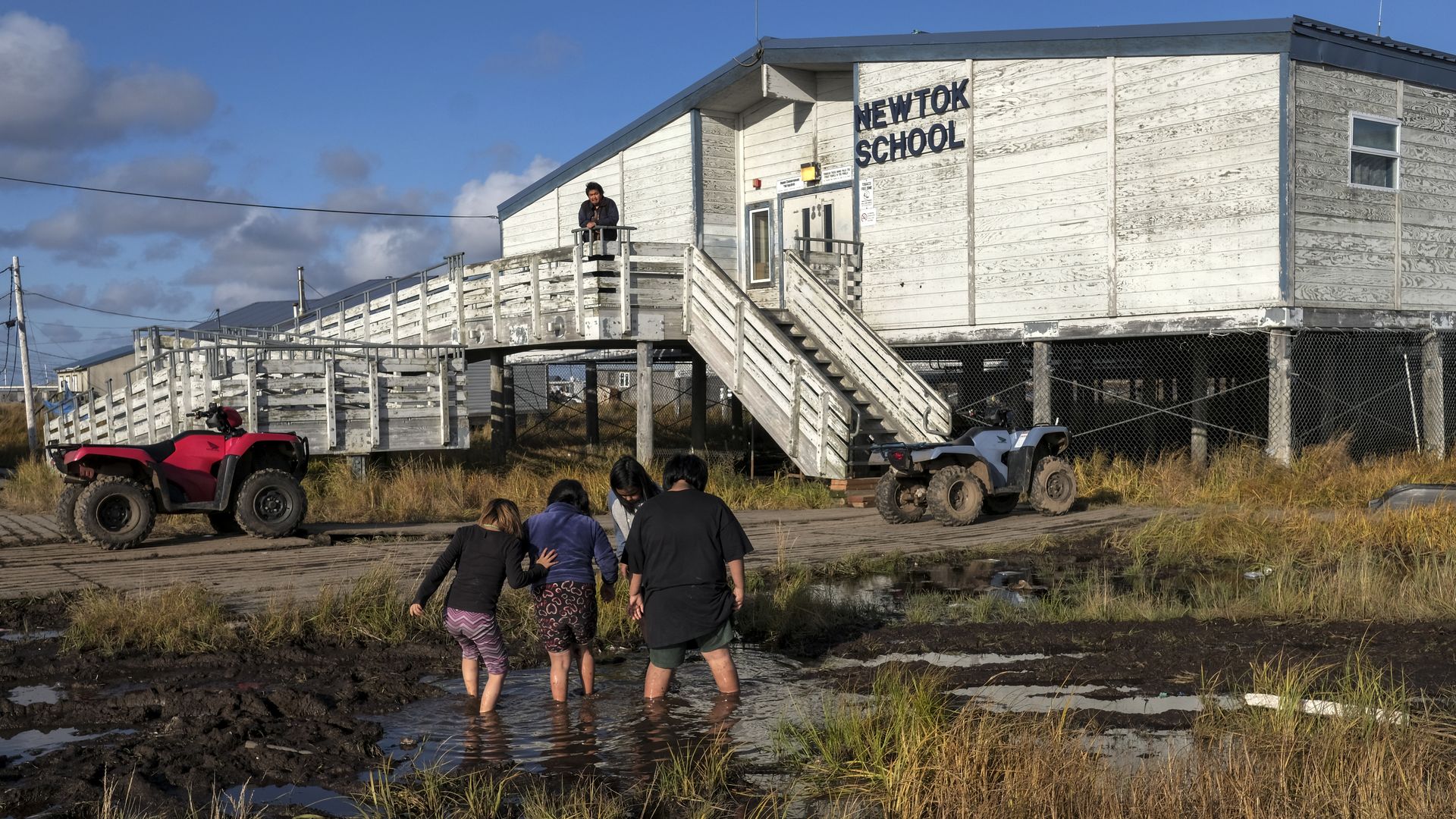 In this image, a group of children stand in the mud outside a white building that says "school"