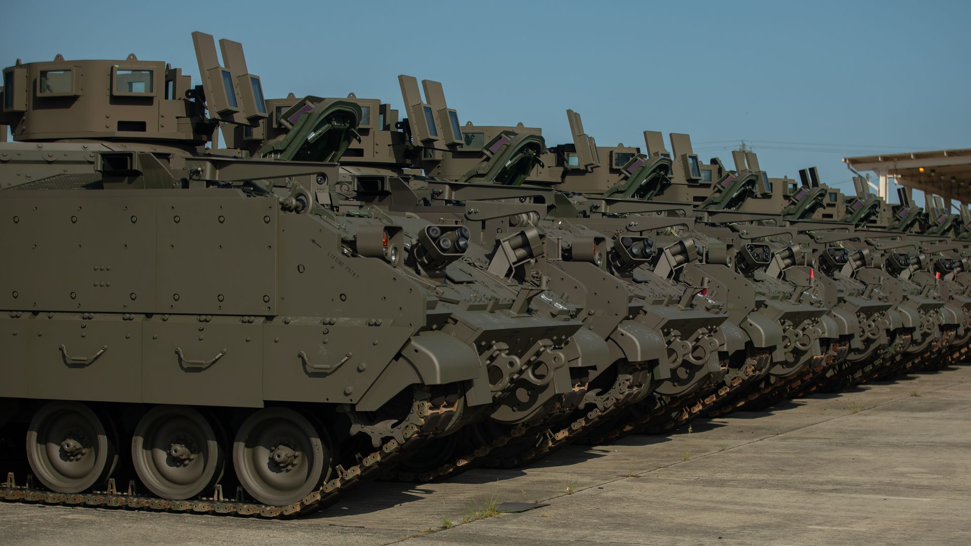 A row of armored vehicles with shielding and tracks visible. They are a greenish-brown color. The sky overhead is clear.