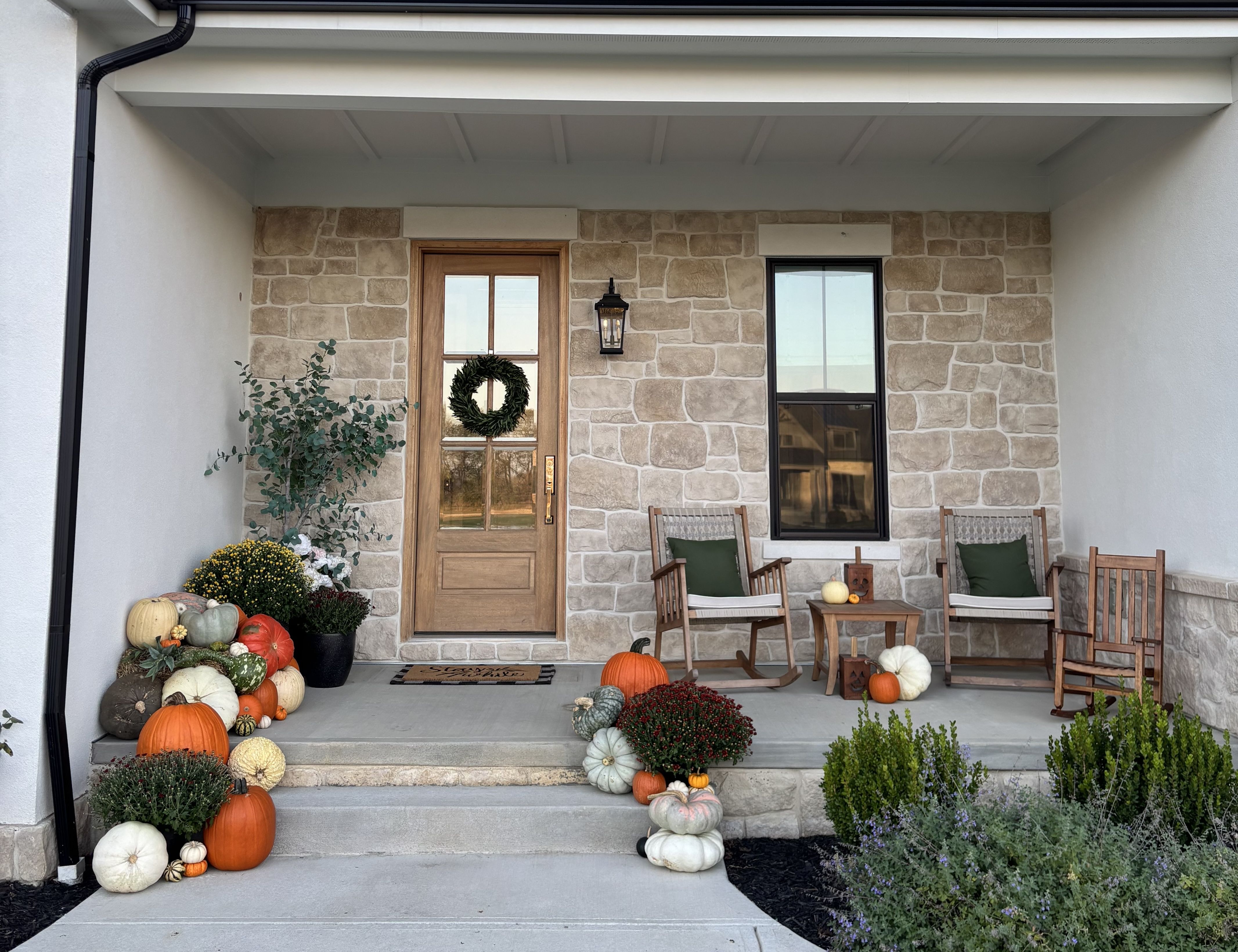 Front porch with beige stone walls, wooden door with green wreath, two wooden rocking chairs with green cushions, table with pumpkins, plants, and autumn decorations including orange and white pumpkins.