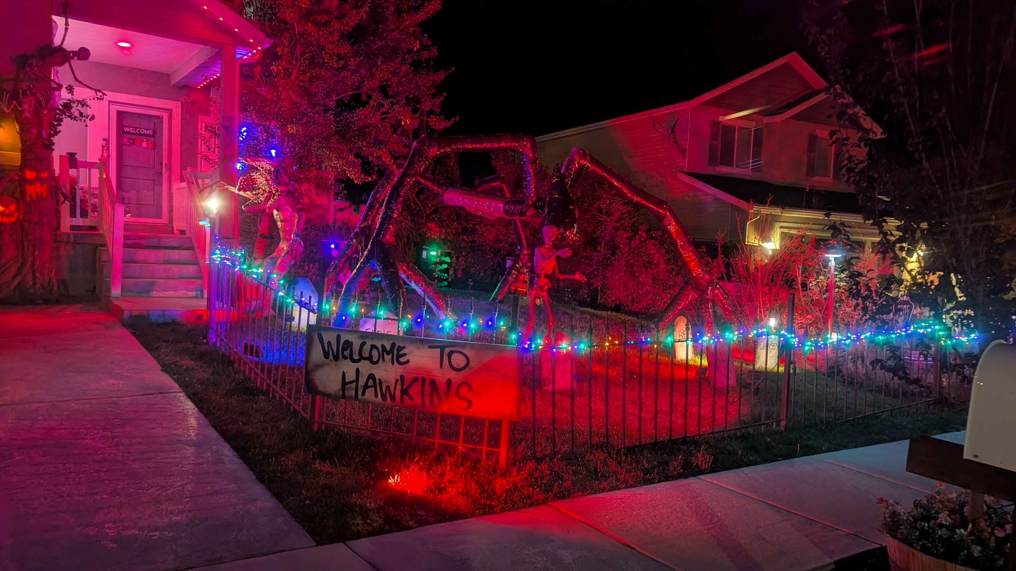 Nighttime Halloween yard with multicolored fence lights, a "WELCOME TO HAWKINS" banner, a carved pumpkin on the porch, and a large red twisted tentacle sculpture near the house.