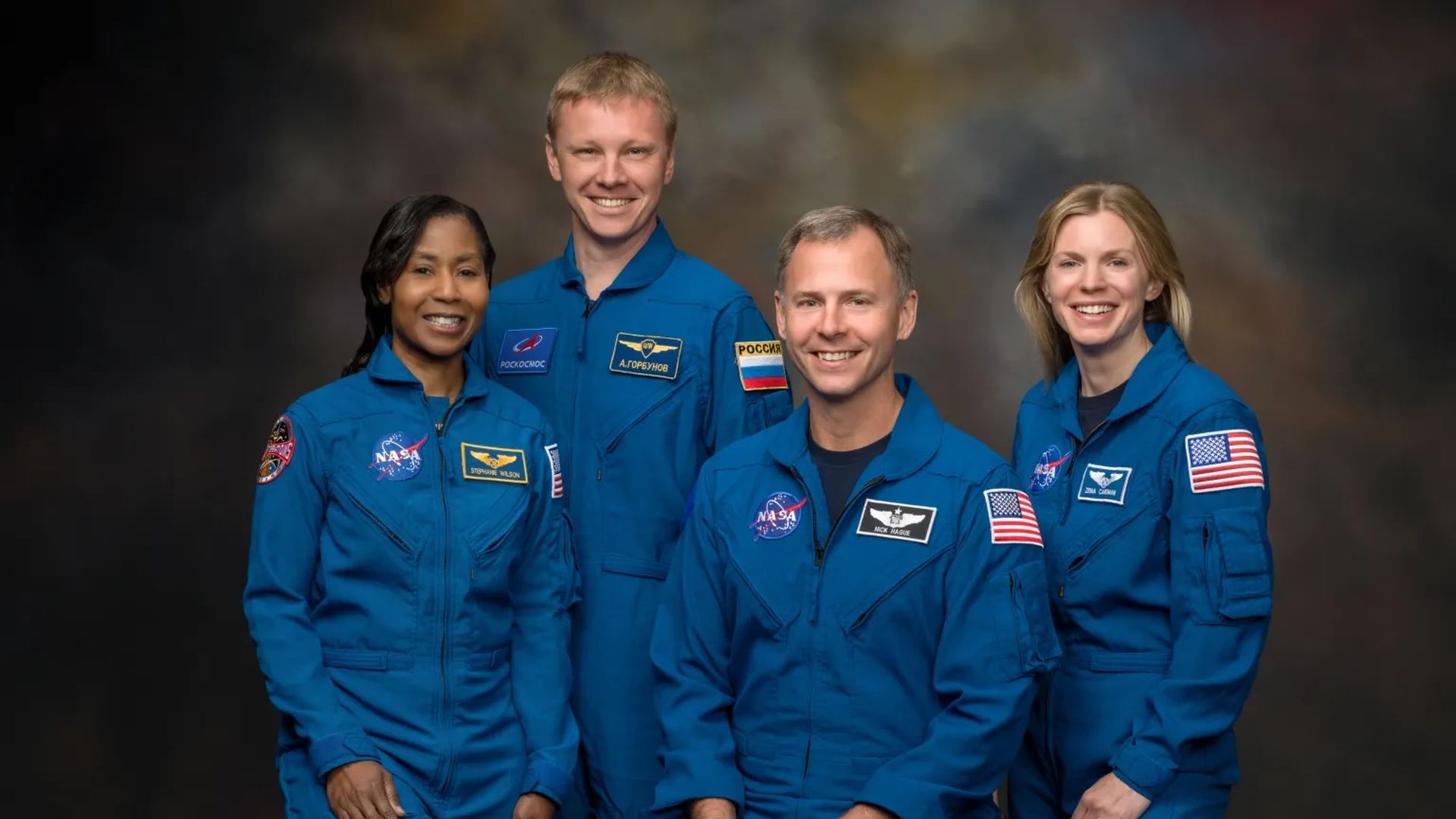 Four astronauts wearing blue jumpsuits pose for a group photo