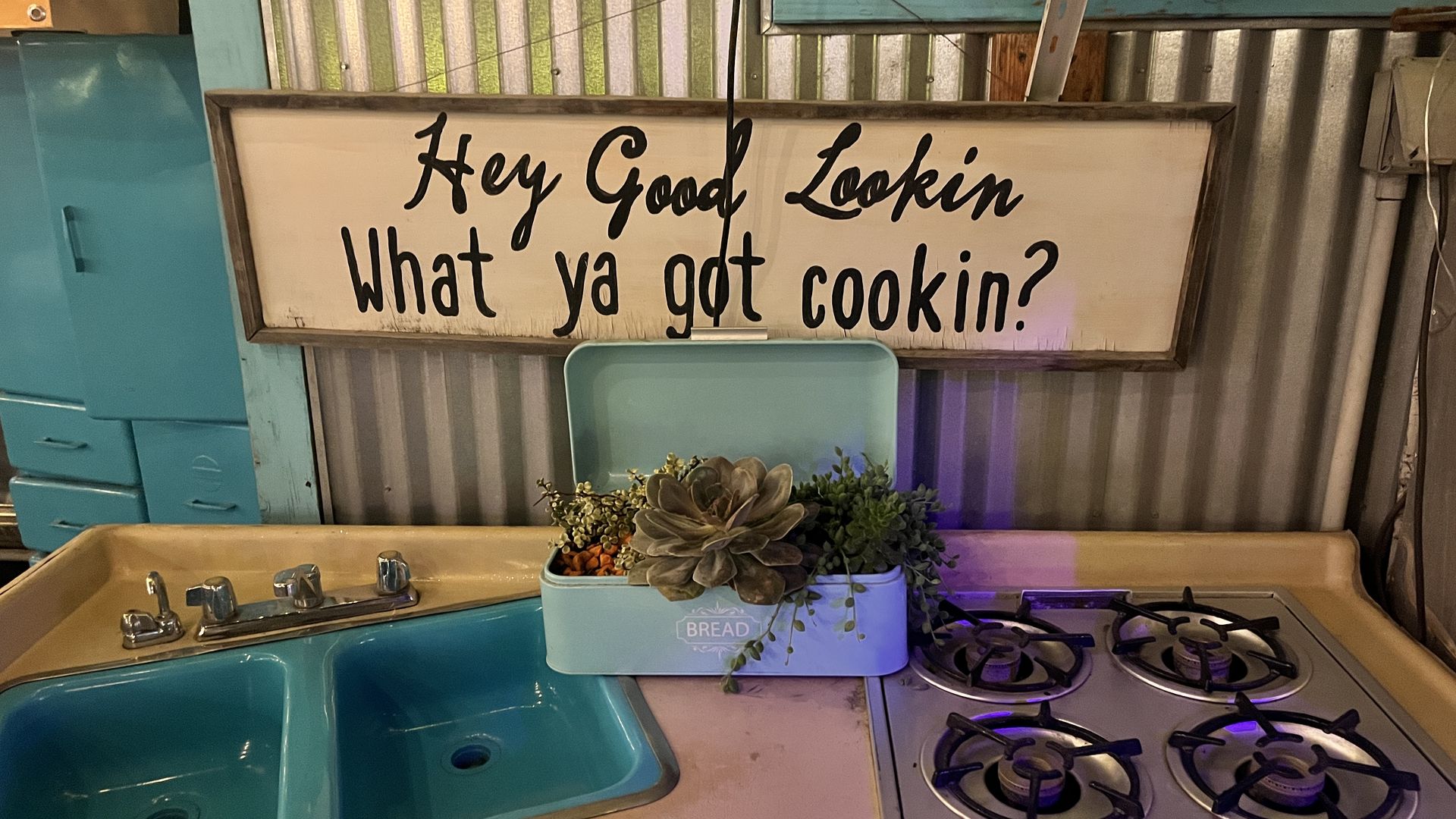 A replica sink and stove with a plant in a box on top of them, all below a sign that reads, "Hey good looking, what ya got cookin?"
